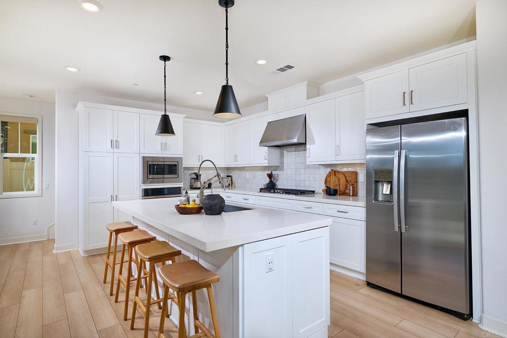 Modern white kitchen with a large island, wooden stools, and stainless steel appliances.