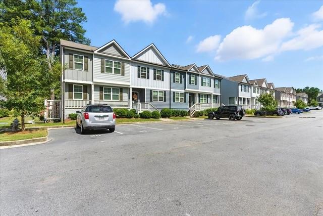 Attached two-story homes with gabled roofs line a paved parking lot beside mature trees.