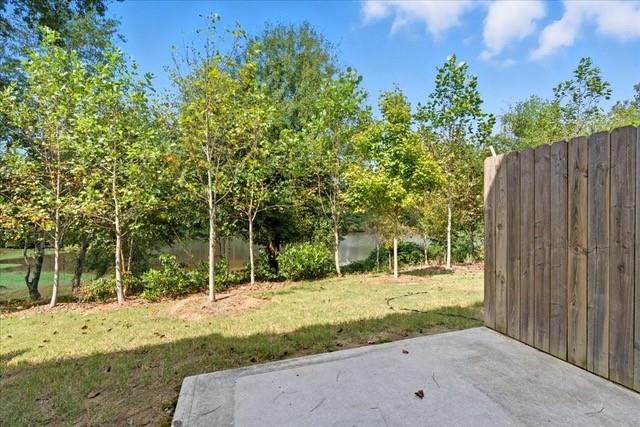 Backyard patio beside a wooden privacy fence, with trees and a glimpse of water beyond the lawn.