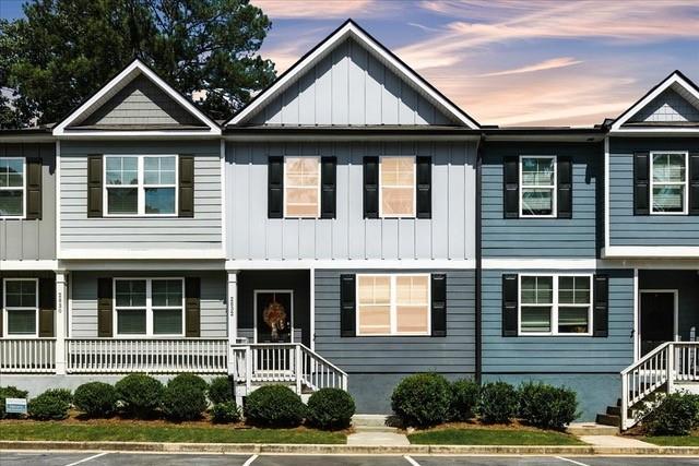 Attached townhomes with gray and blue siding, front porches, and shrubs beneath a pink evening sky.