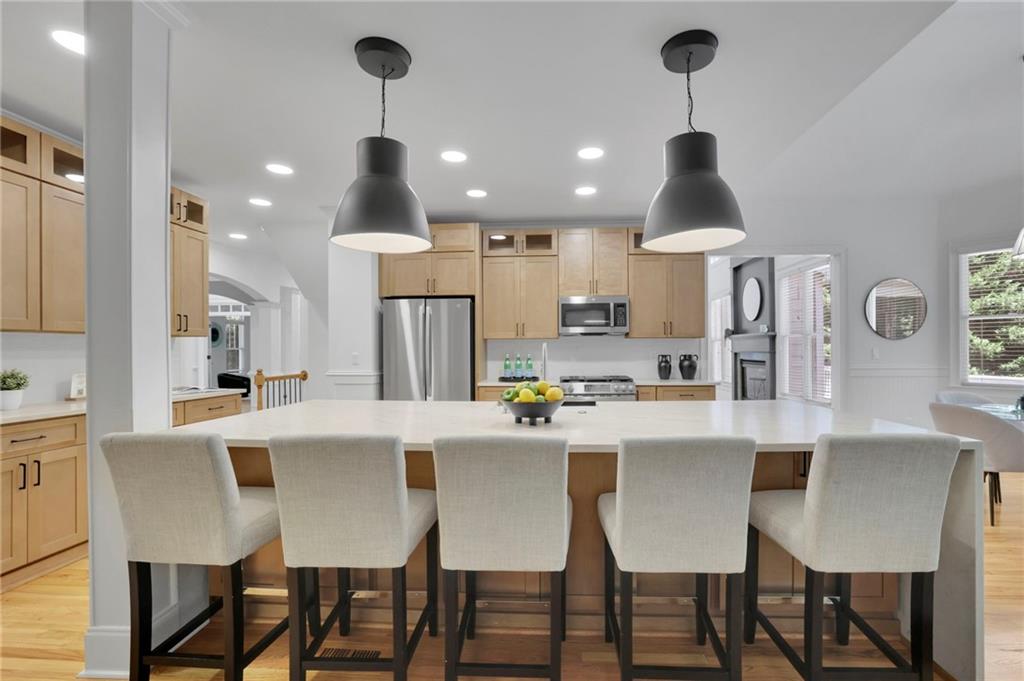 Kitchen with a large white island, five upholstered stools, light wood cabinets, and stainless steel appliances.