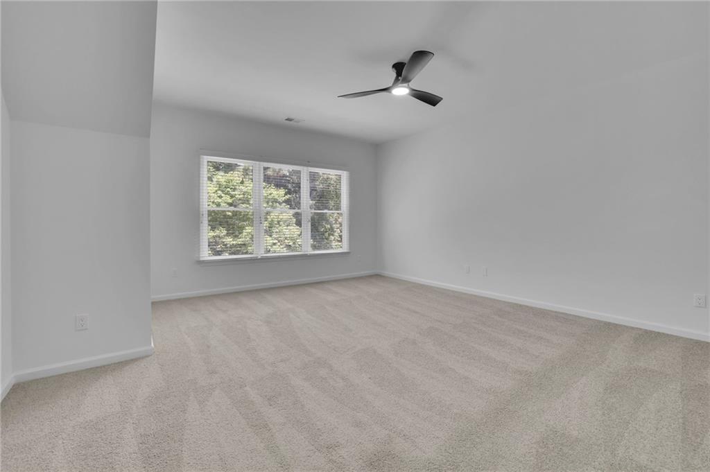 Empty carpeted room with a wide window overlooking trees and a ceiling fan with light.