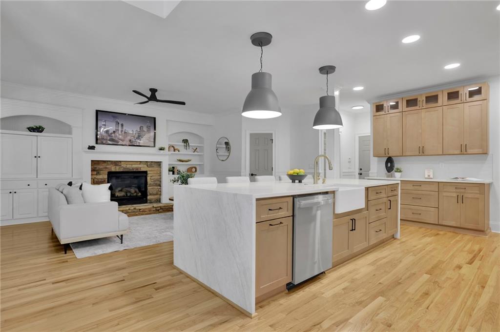 Open kitchen with a large white island and light wood cabinets beside a living area with a stone fireplace.