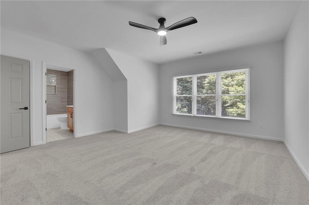 Empty carpeted bedroom with a ceiling fan, wide window, and adjoining bathroom.