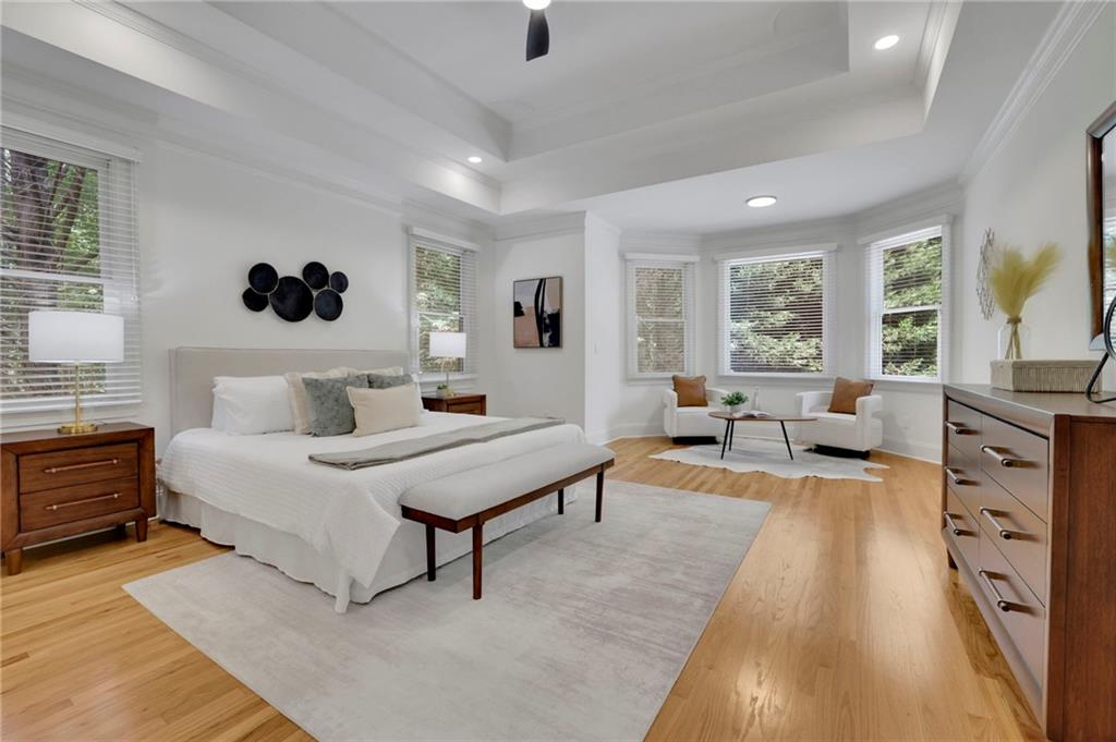 Staged bedroom with a white bed, tray ceiling, hardwood floors, and a bay-window sitting area.