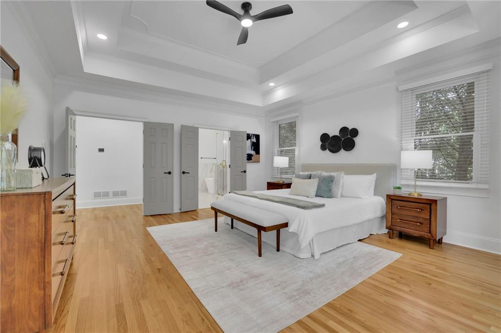 Bright staged bedroom with a white bed, hardwood floors, large windows, and a tray ceiling.