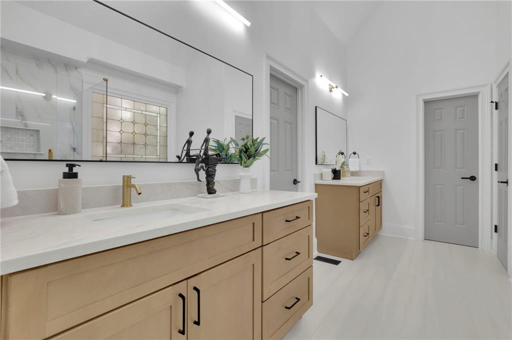 Modern bathroom with a long light-wood vanity, white counters, gold faucets, and a glass shower reflected in the mirror.
