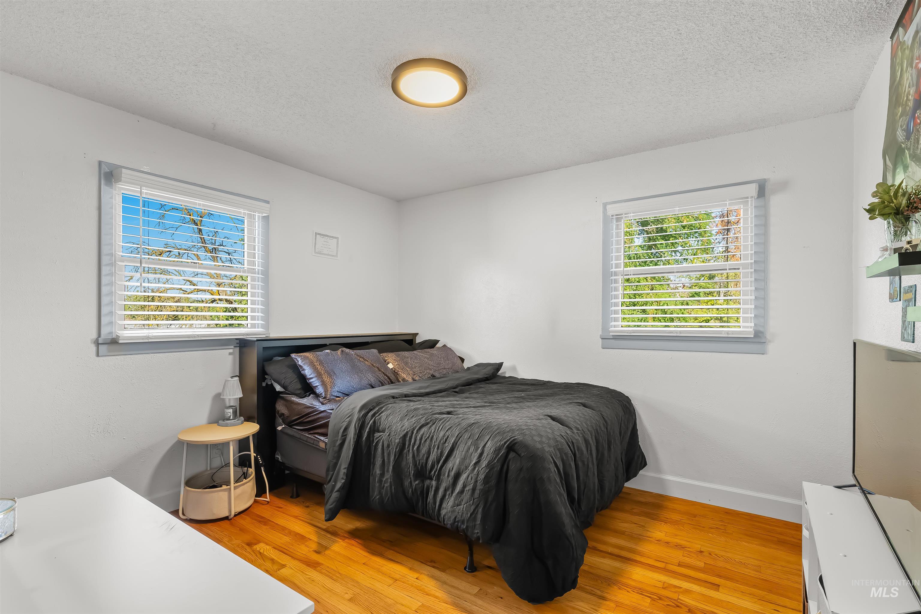 Furnished bedroom with a dark bed, two bright windows, and warm hardwood floors.