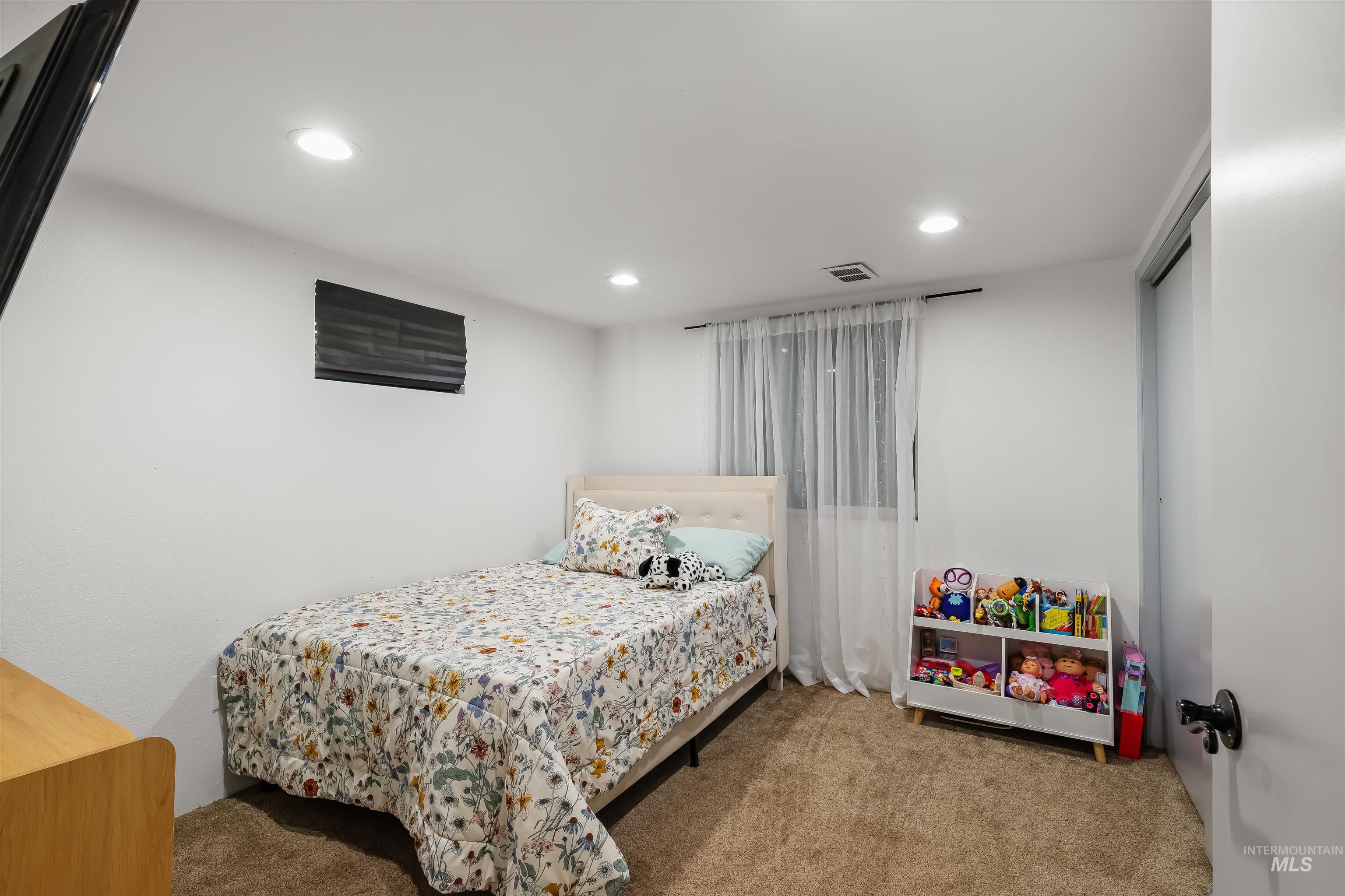 Bedroom with floral bedding, beige carpet, and a toy shelf beside the bed.