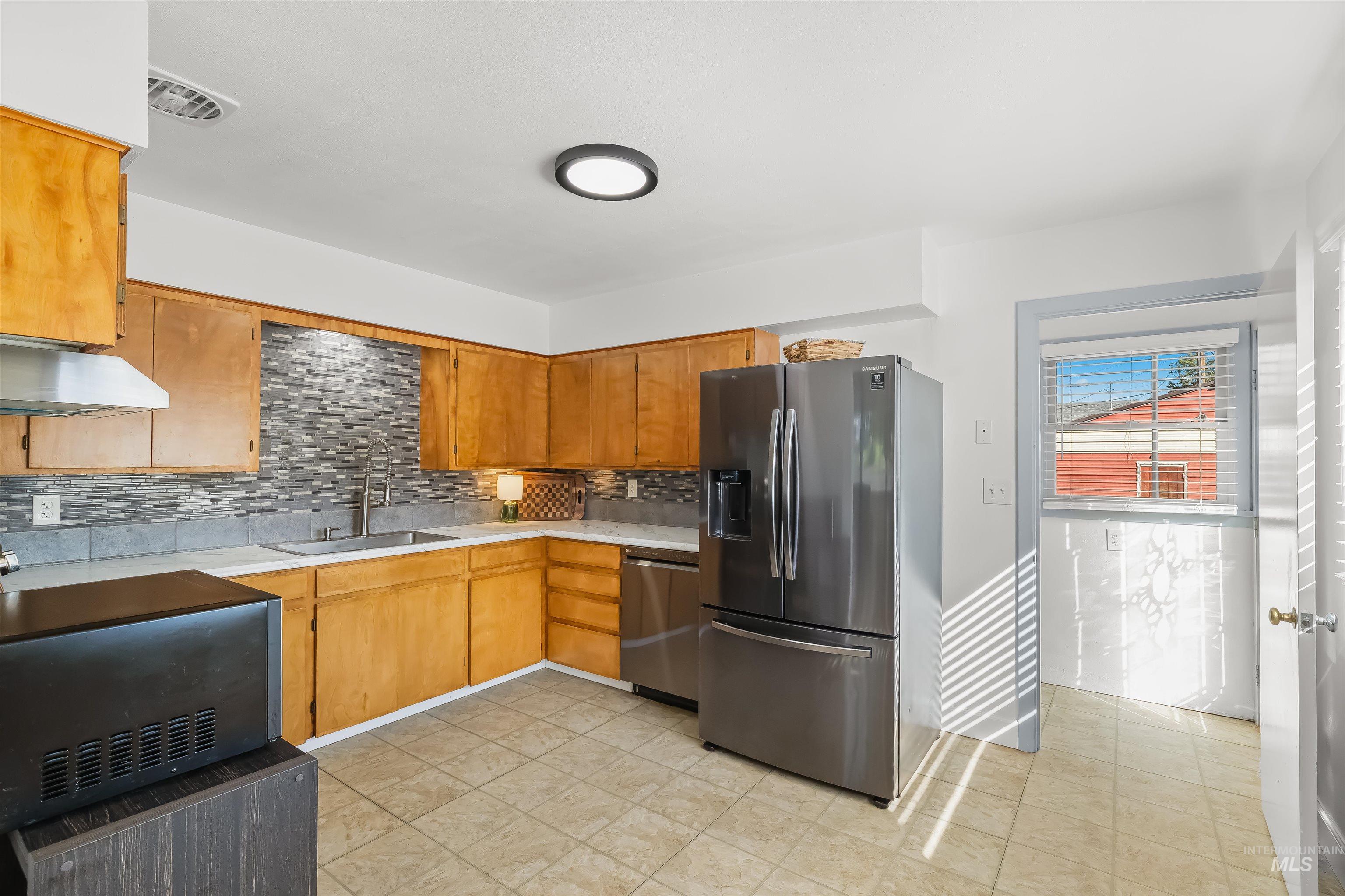 Kitchen with honey-colored cabinets, gray mosaic backsplash, and a large stainless steel refrigerator.