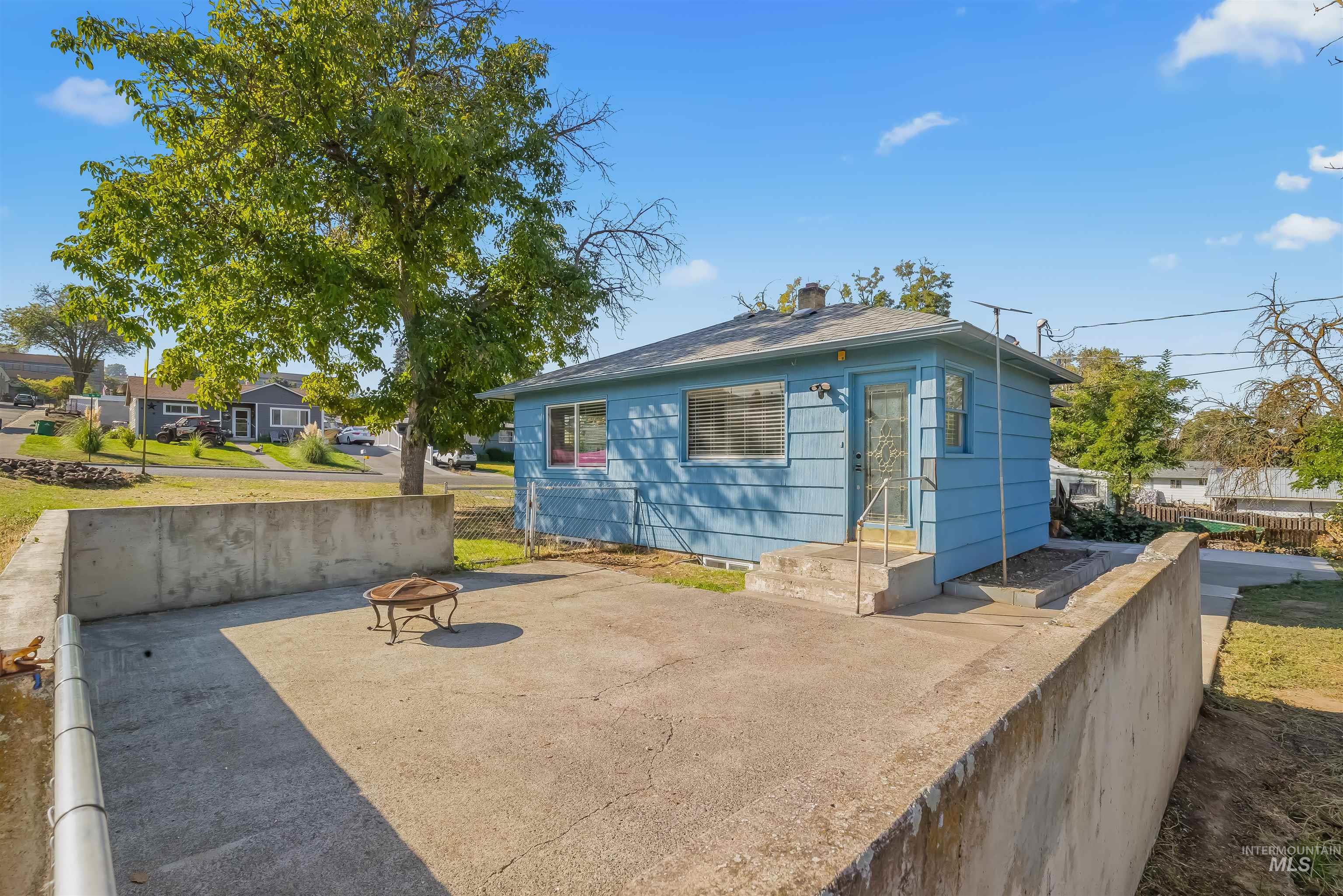 Blue-sided house with a raised concrete patio, low walls, and a small metal fire pit.