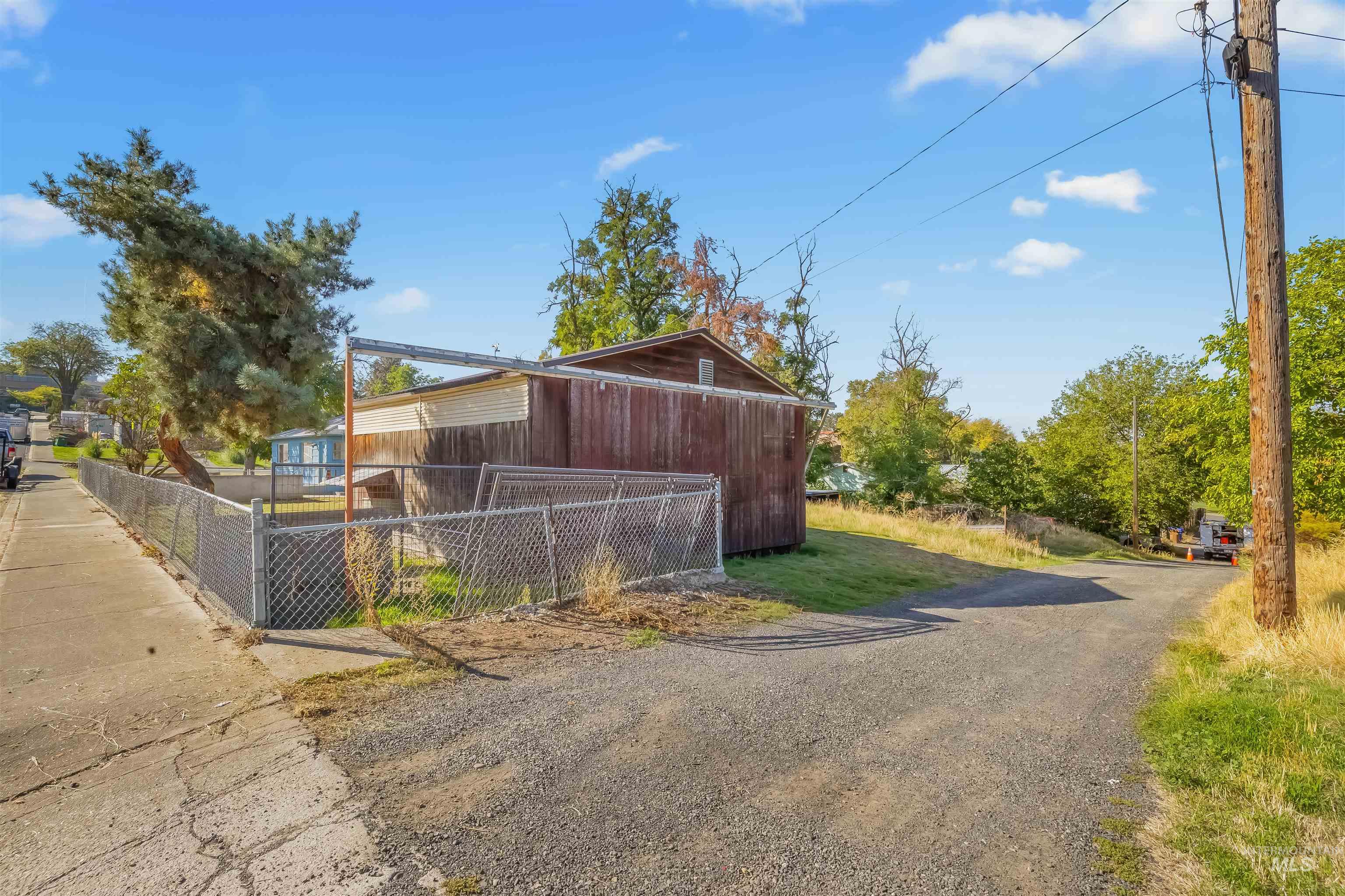 Weathered brown outbuilding behind chain-link fencing beside a gravel driveway.