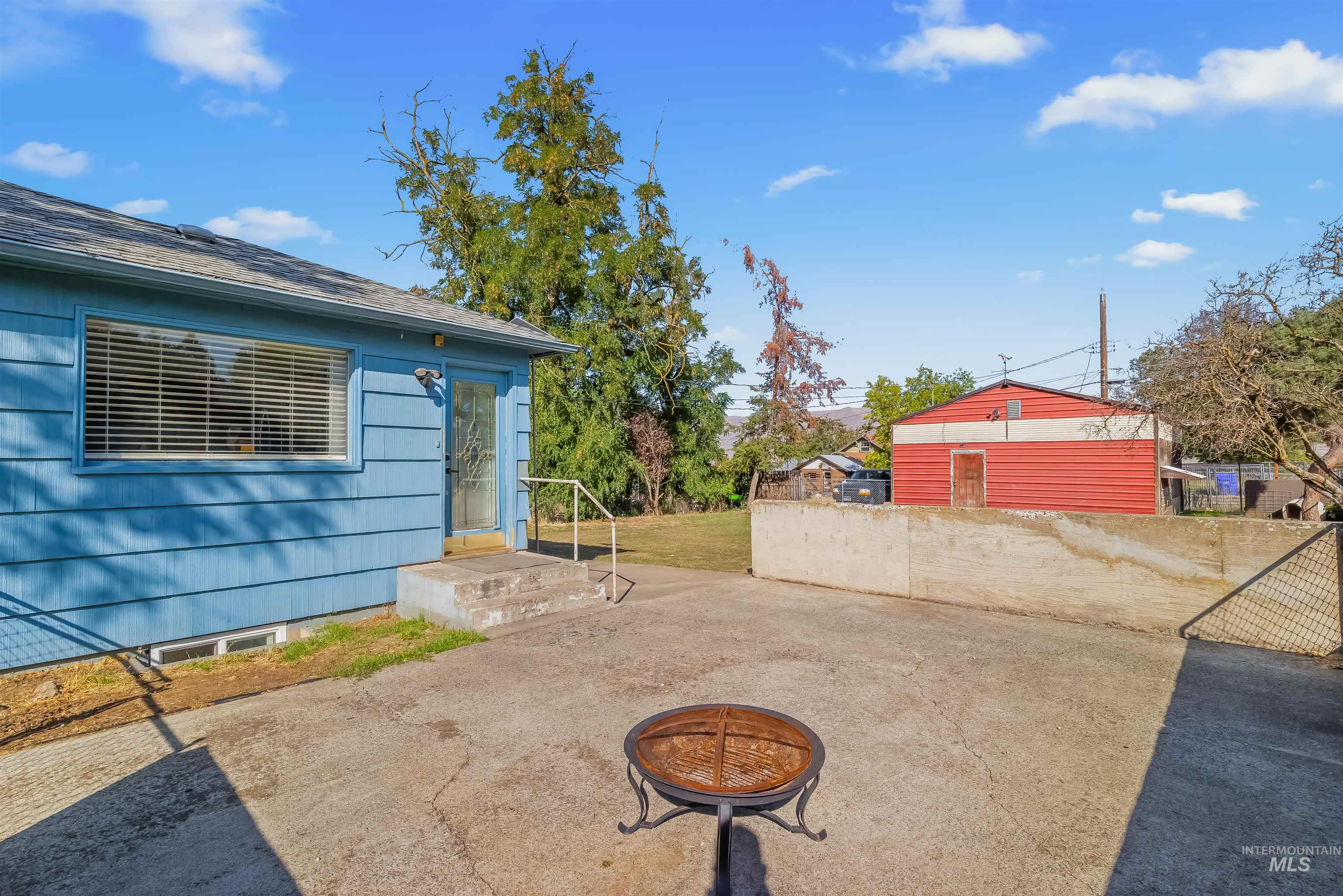 Blue-sided house beside a paved patio with a metal fire pit and red outbuilding.