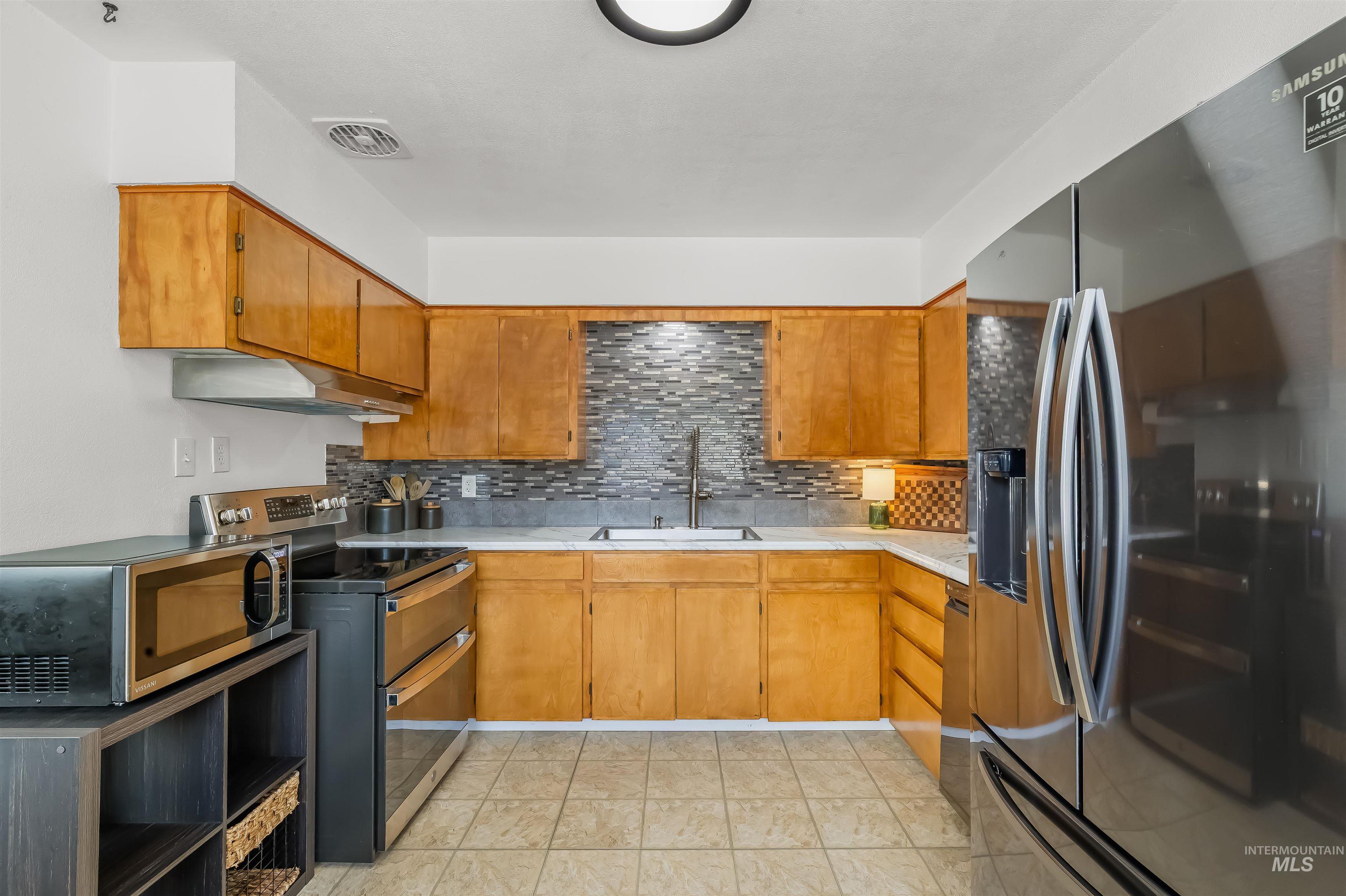 Kitchen with honey-colored cabinets, gray mosaic backsplash, and a large stainless-steel refrigerator.