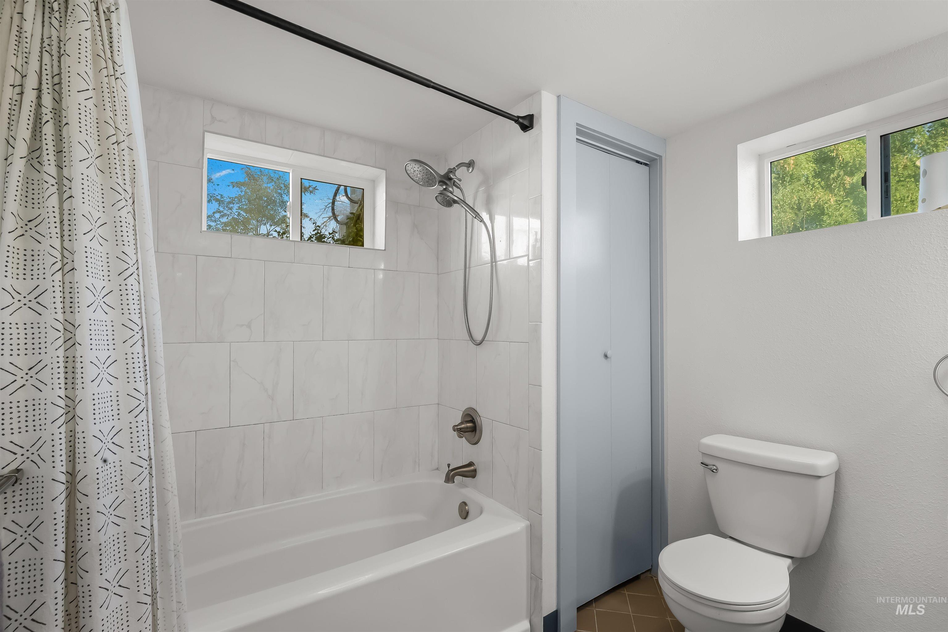 Bathroom with marble-look tub surround, patterned shower curtain, blue-gray storage door, and high window.