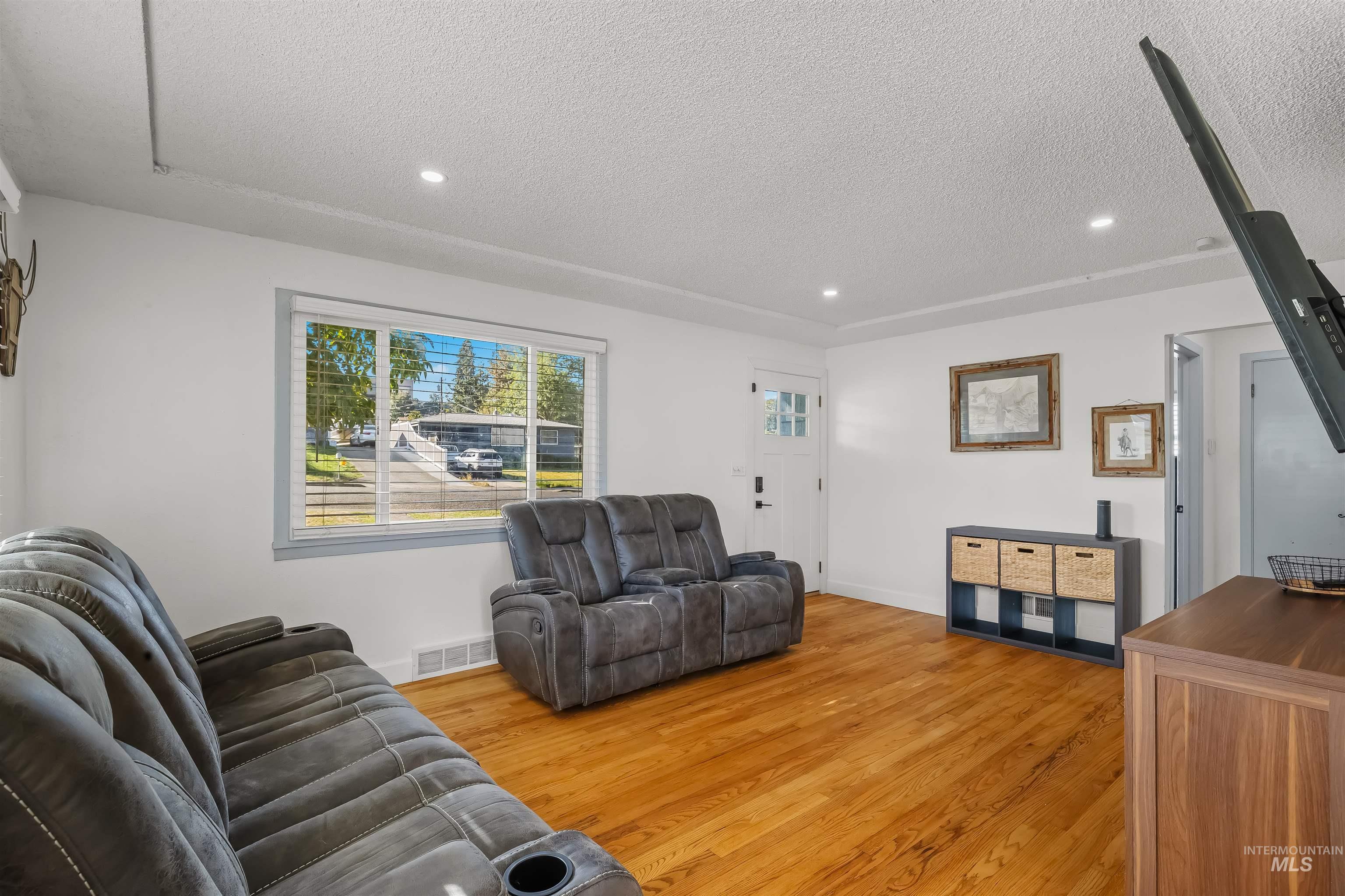 Living room with gray reclining sofas, hardwood floors, and a large window overlooking the street.