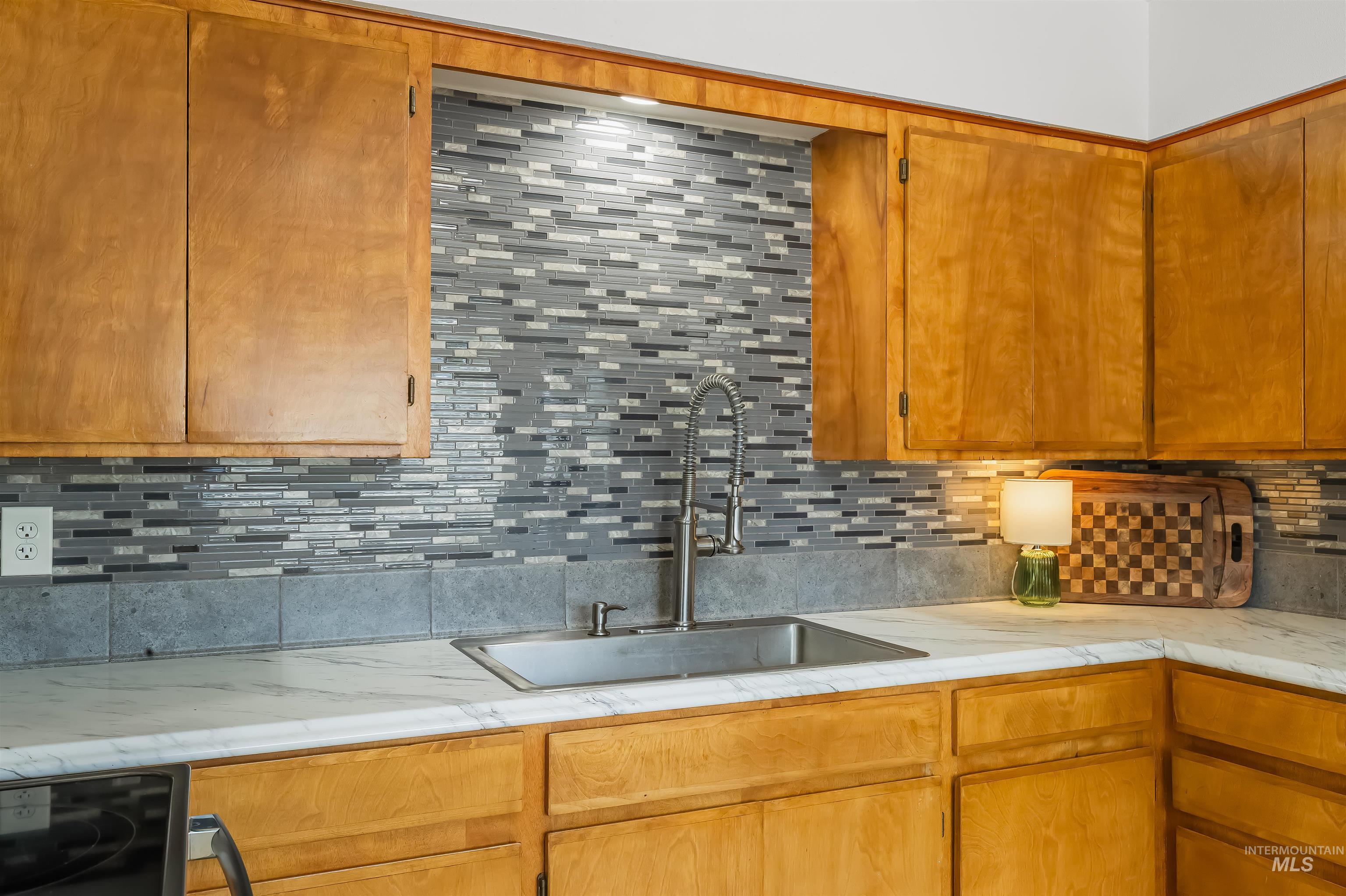 Kitchen sink beneath honey-colored cabinets, with a gray tile backsplash and white marble-look counters.