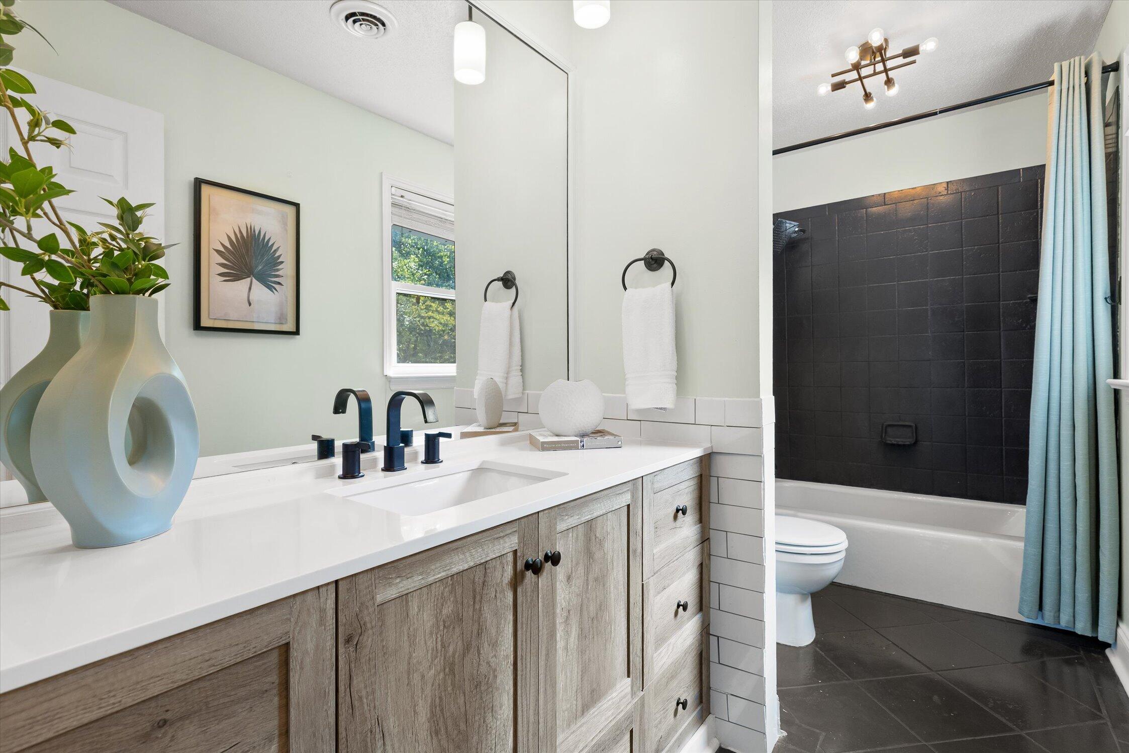 Bright bathroom with a wood vanity and white counter, alongside a black-tiled tub and pale blue shower curtain.