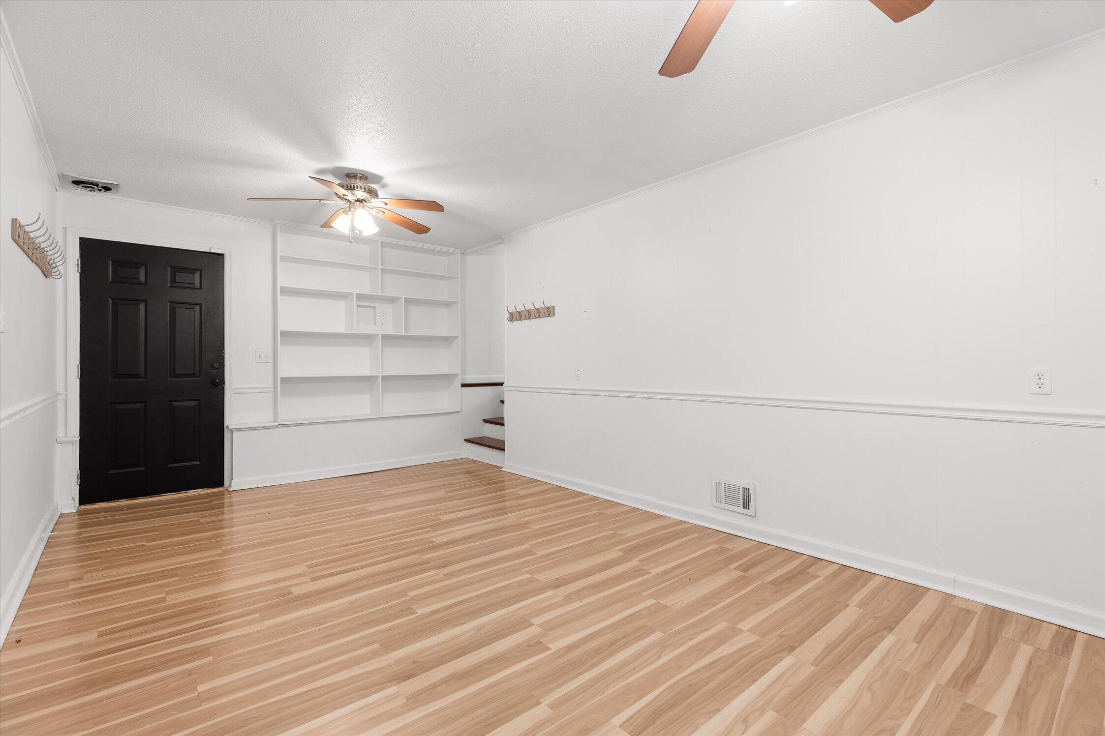 Empty room with light wood floors, built-in white shelves, a black door, and a ceiling fan.