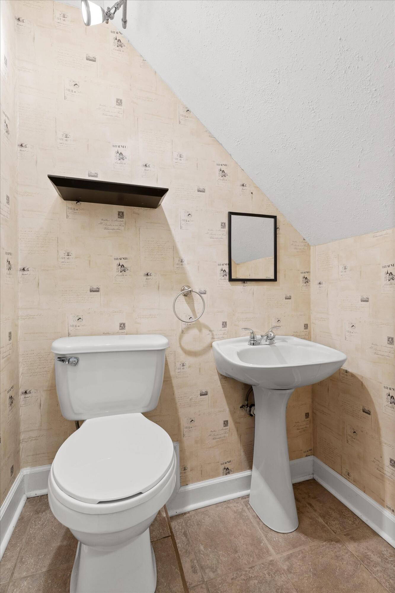 Bathroom with a white toilet and pedestal sink beneath a sloped ceiling, with beige patterned wallpaper.