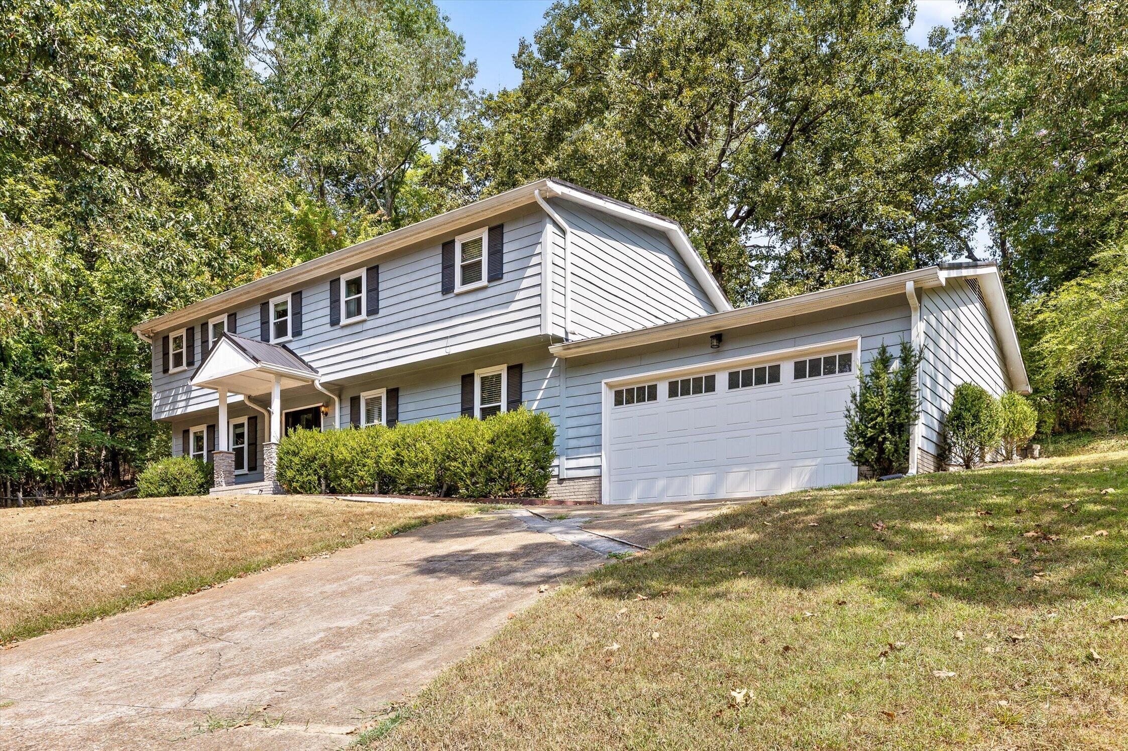 Light blue two-story house with black shutters, a covered entry, and an attached garage amid mature trees.