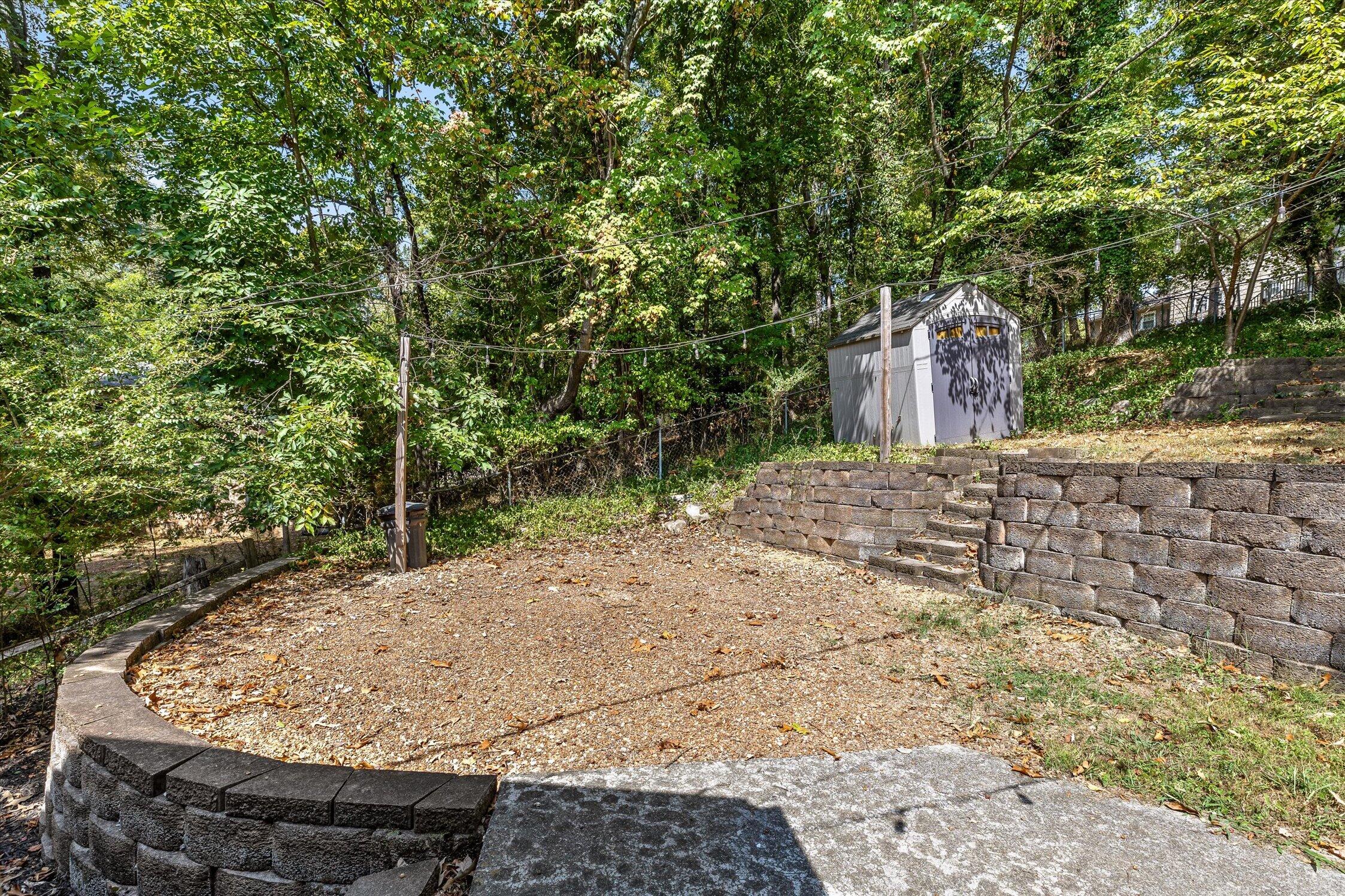 Wooded backyard with a gravel area, curved stone retaining walls, steps, and a gray storage shed.