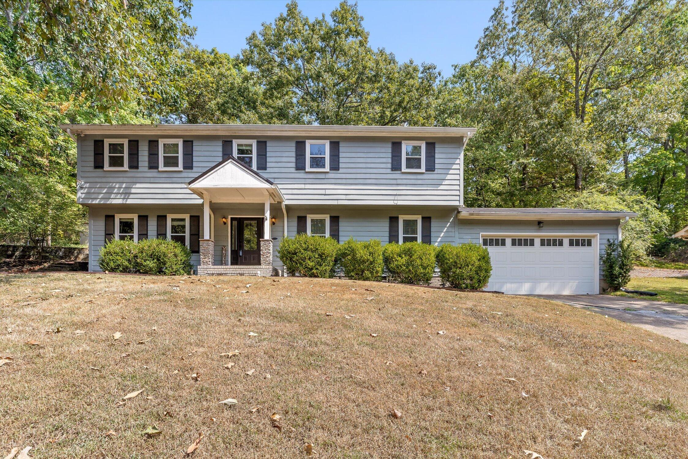 Two-story blue-gray house with a covered entry, attached garage, and mature trees.