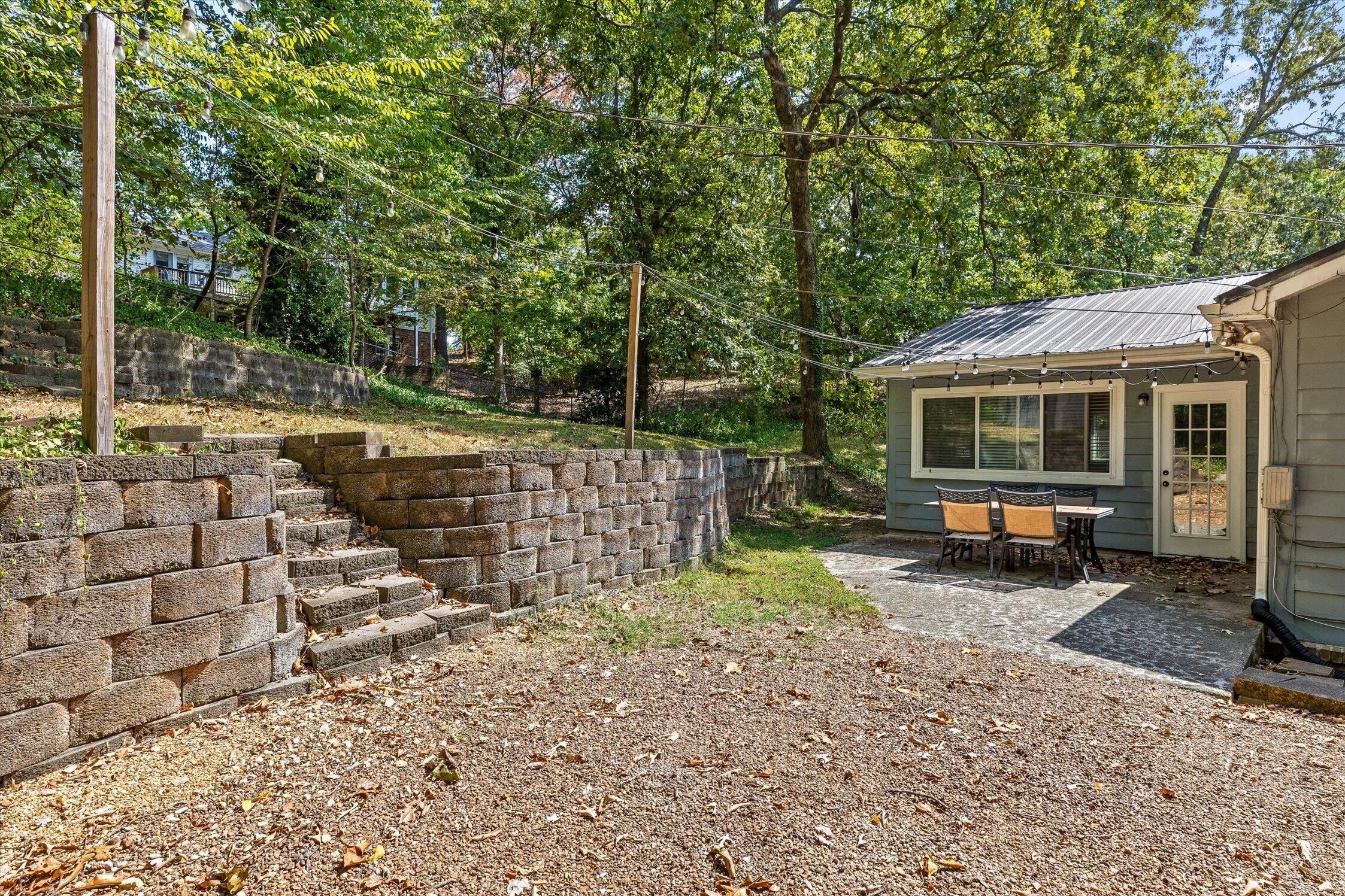 Backyard patio with outdoor dining set, gravel ground, and tiered stone retaining walls beneath mature trees.