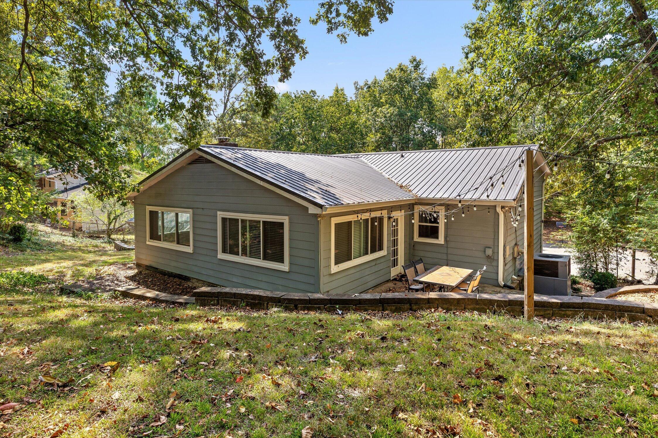 Gray single-story home with a metal roof and string-lit patio beneath mature trees.