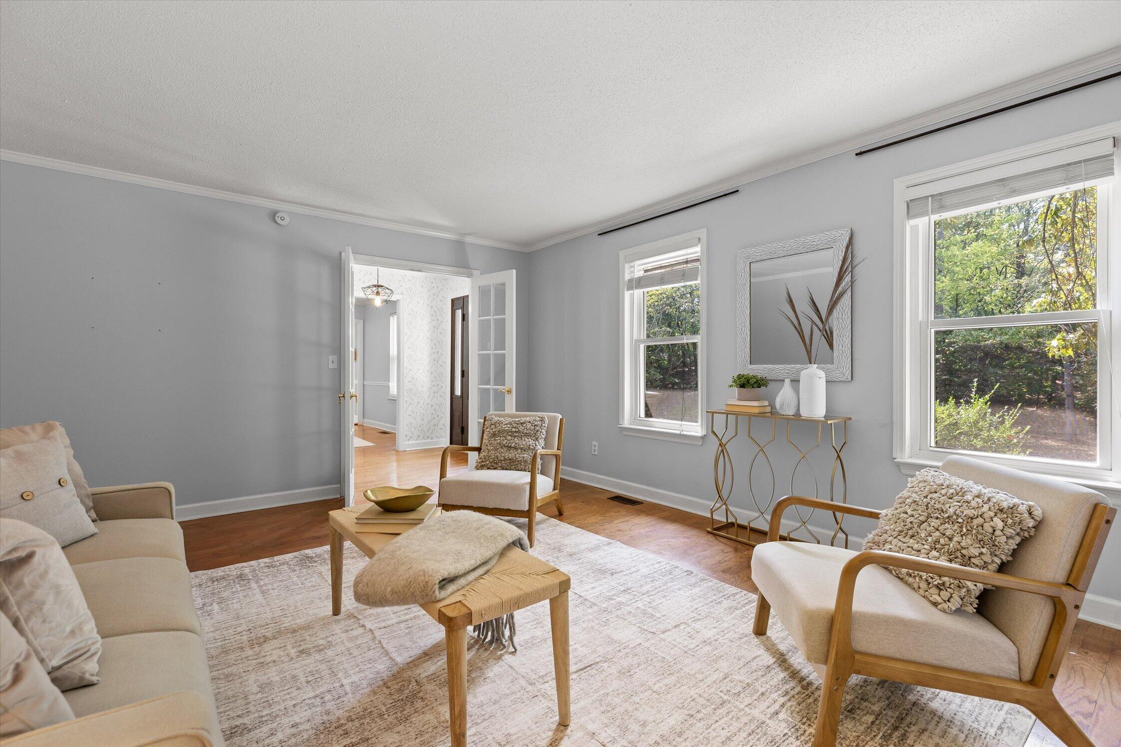 Staged living room with neutral seating, hardwood floors, and windows overlooking trees.