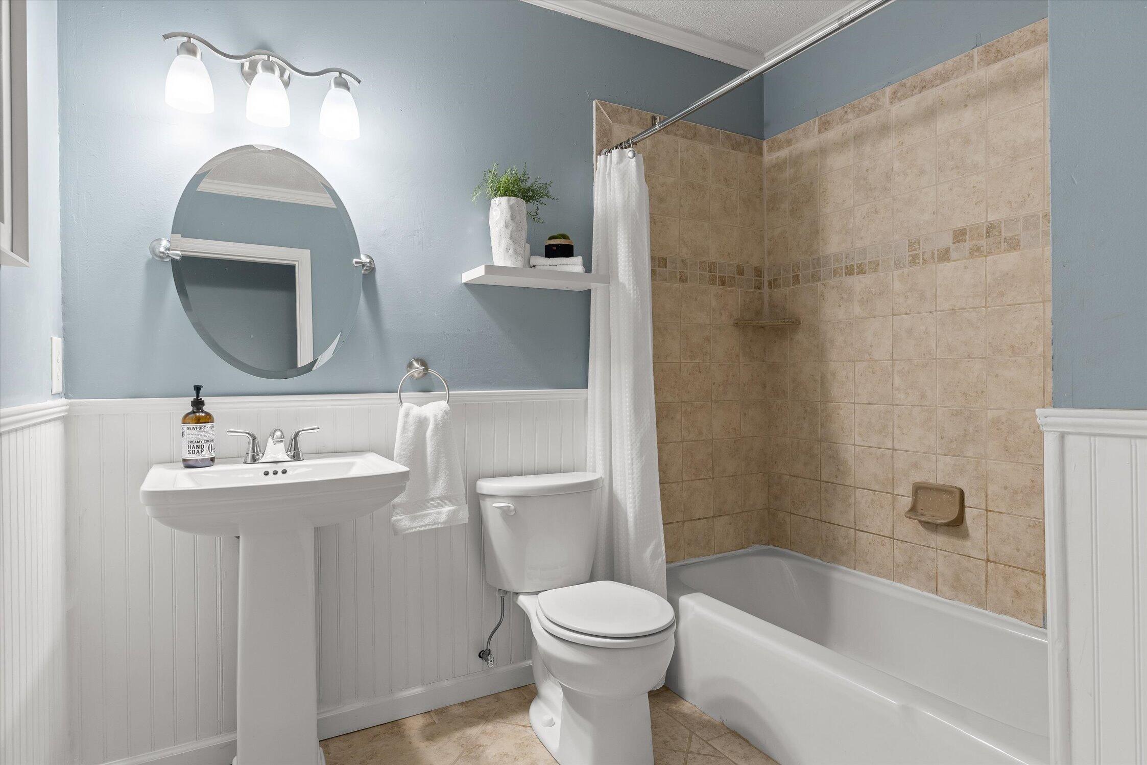 Bathroom with a pedestal sink, round mirror, and bathtub lined with tan tile.