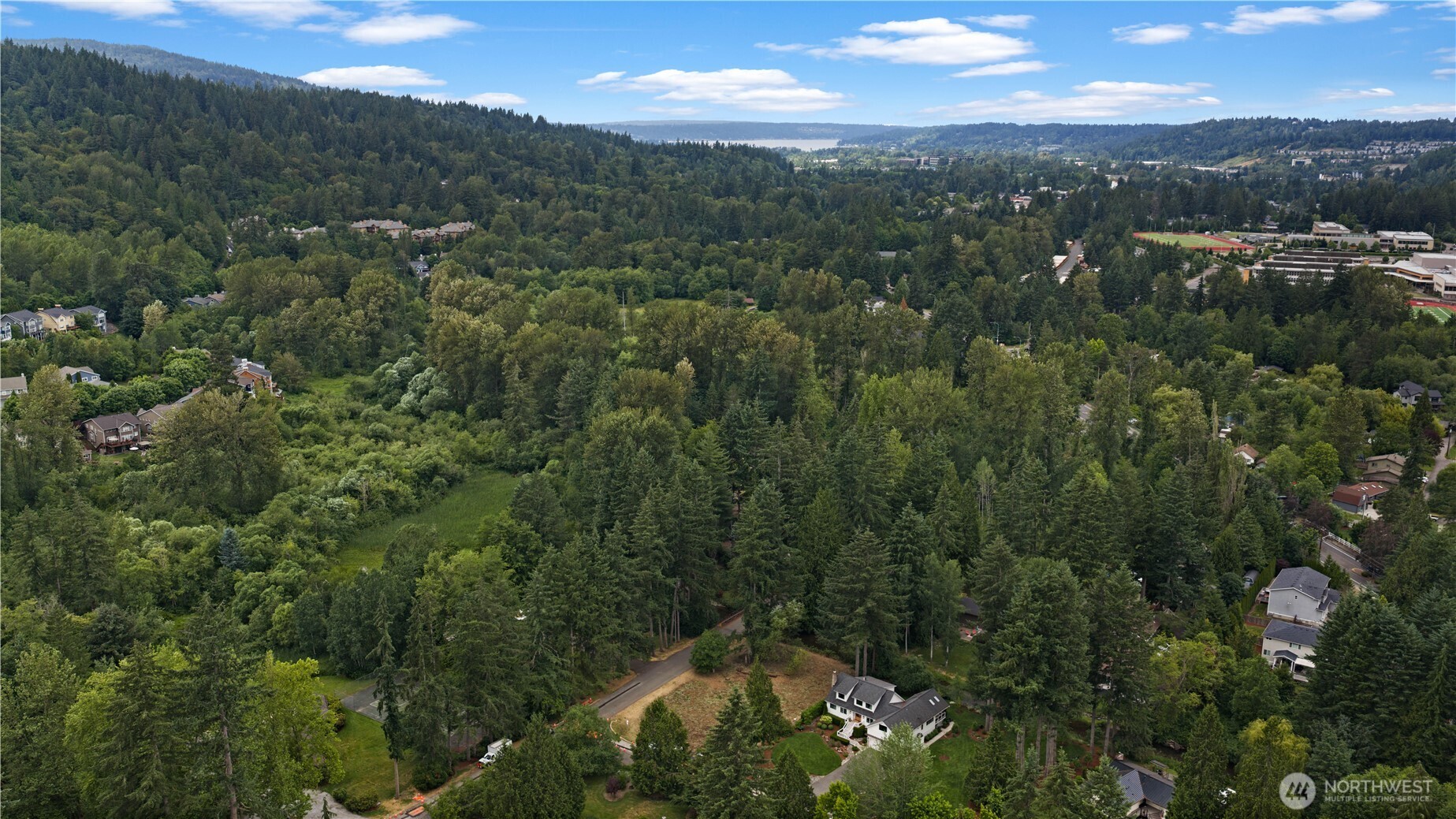 Aerial view of a wooded neighborhood with scattered homes, forested hills, and distant water.