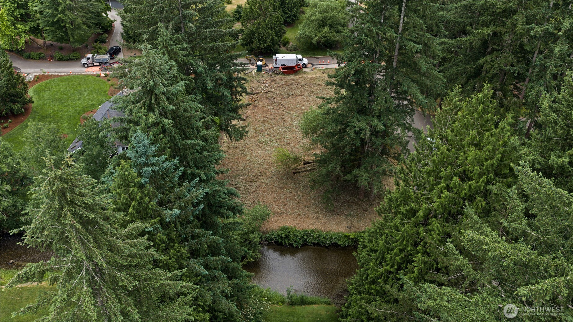 Aerial view of a cleared, wooded property beside a creek, with work trucks on the road.