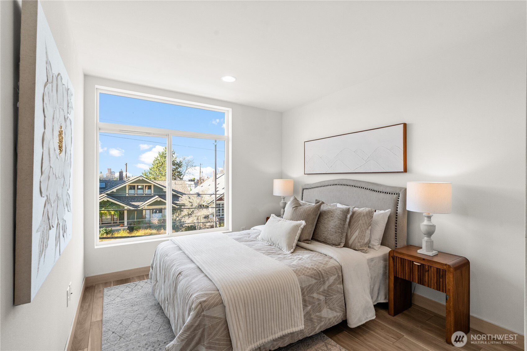 Staged bedroom with a gray upholstered bed and a large window overlooking nearby homes.