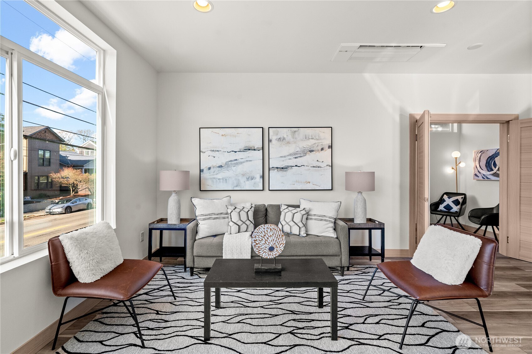 Staged living room with a gray sofa, leather chairs, abstract art, and a large street-facing window.