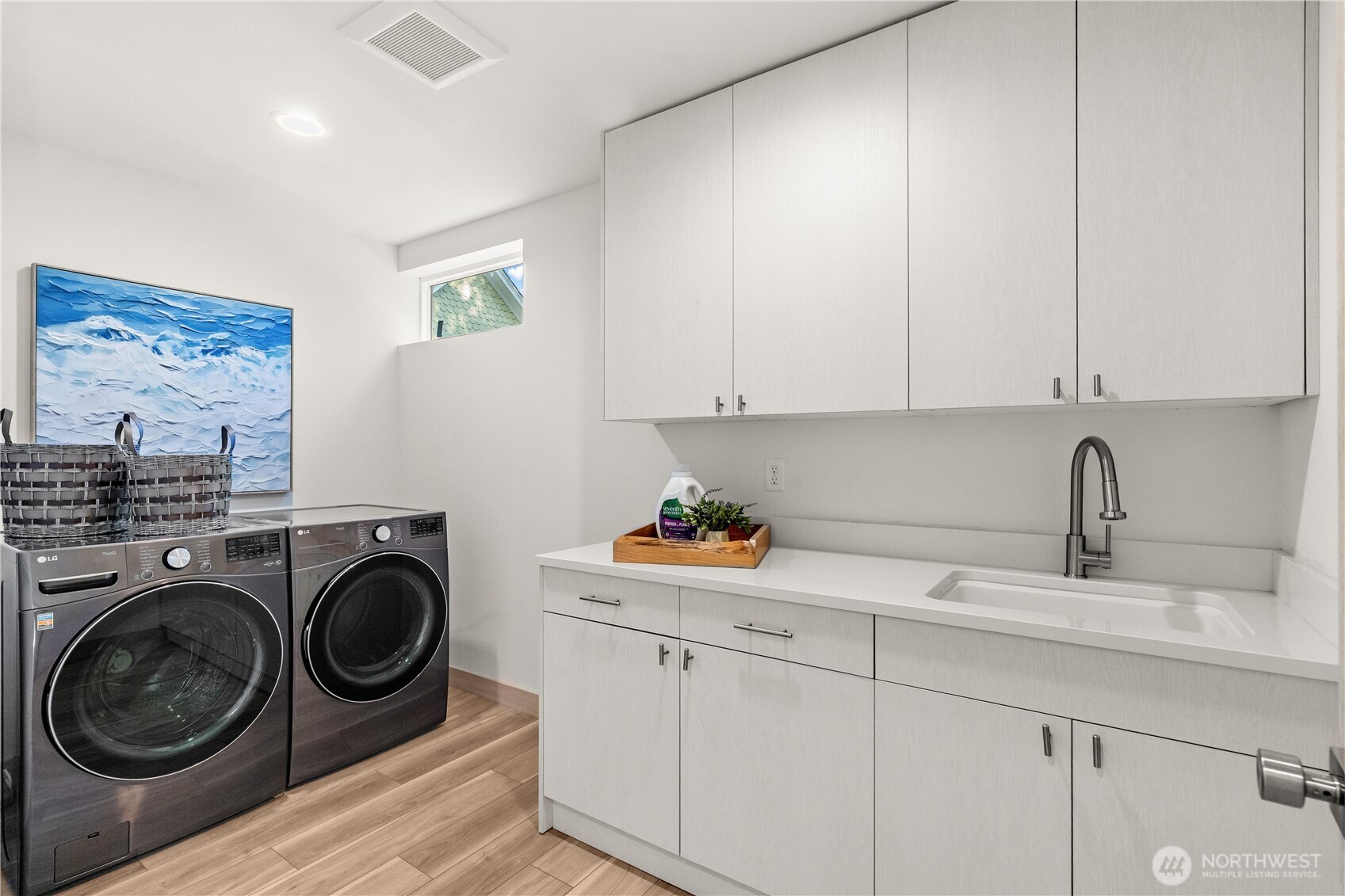 Laundry room with dark front-loading washer and dryer, white cabinetry, and a countertop utility sink.