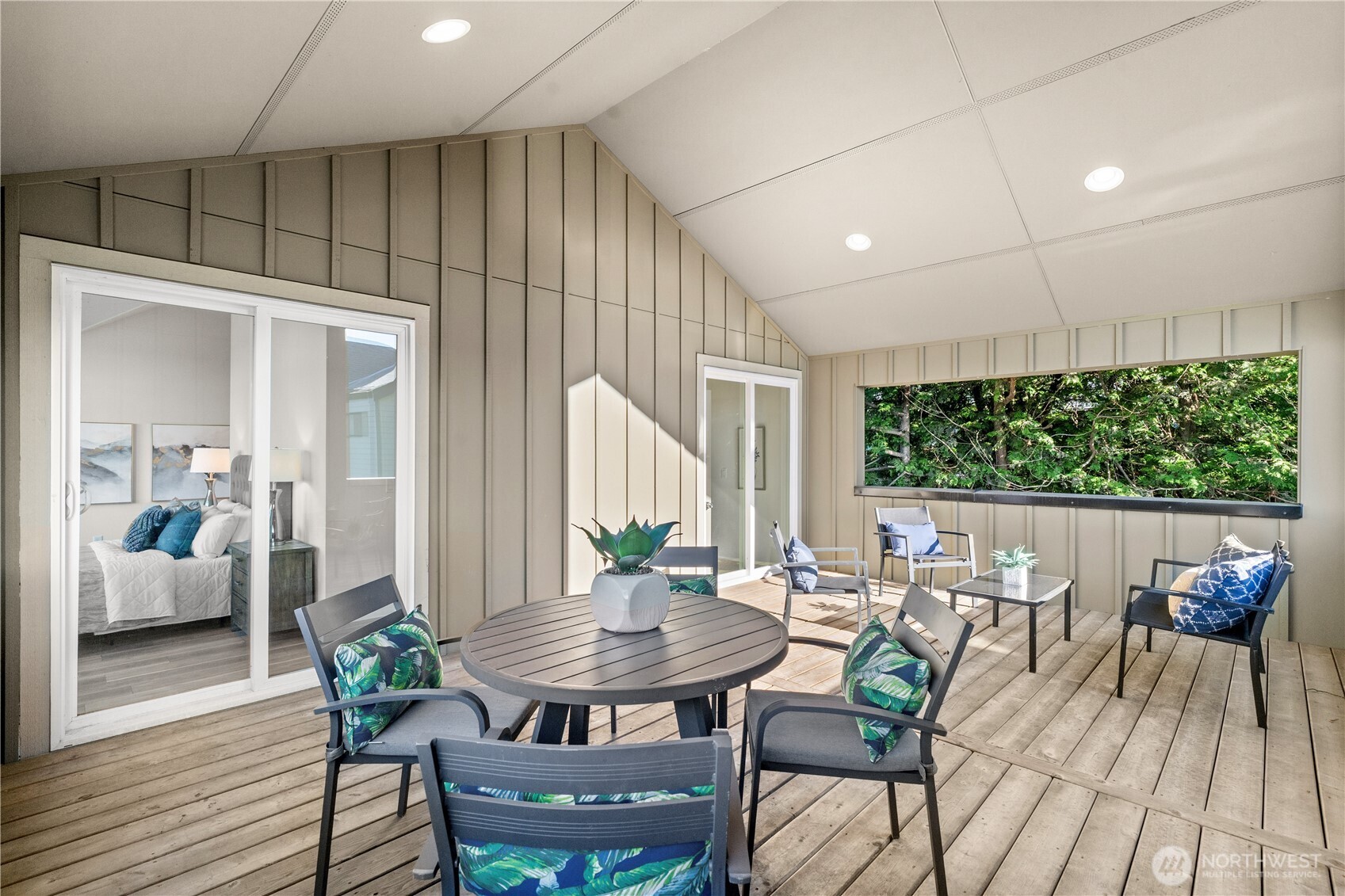 Covered wooden deck with patio seating, sliding doors to a bedroom, and a window facing leafy trees.