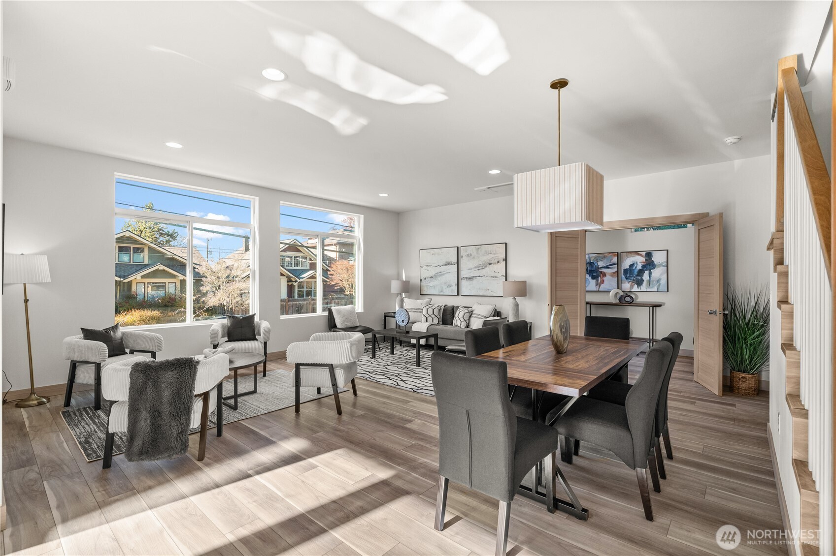 Open-plan living and dining room with large windows, staged seating, and a wood dining table.