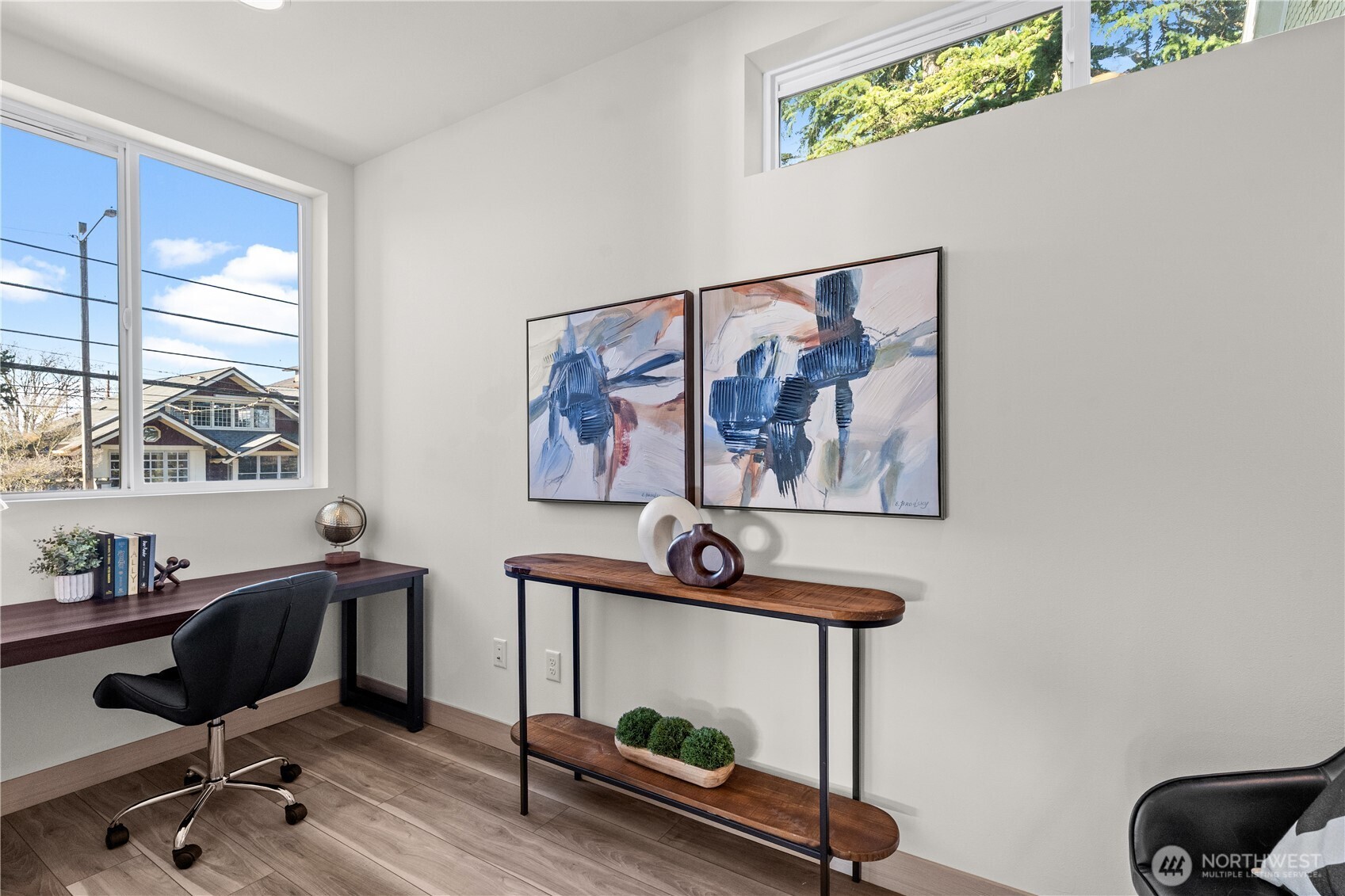 Staged home office with a desk, large windows, abstract artwork, and a wood console table.
