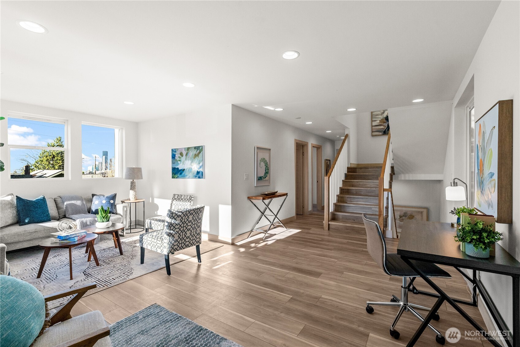Staged living and dining area with gray seating, wood-look floors, and a staircase; Seattle skyline visible through the windows.