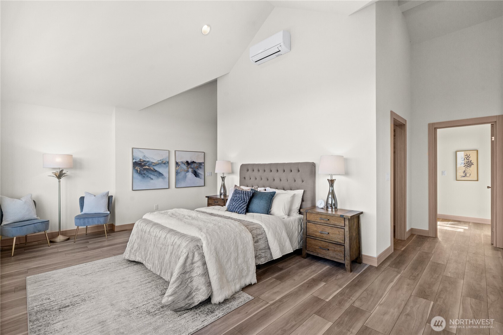 Staged bedroom with a tufted gray headboard, blue accent chairs, and vaulted ceiling.