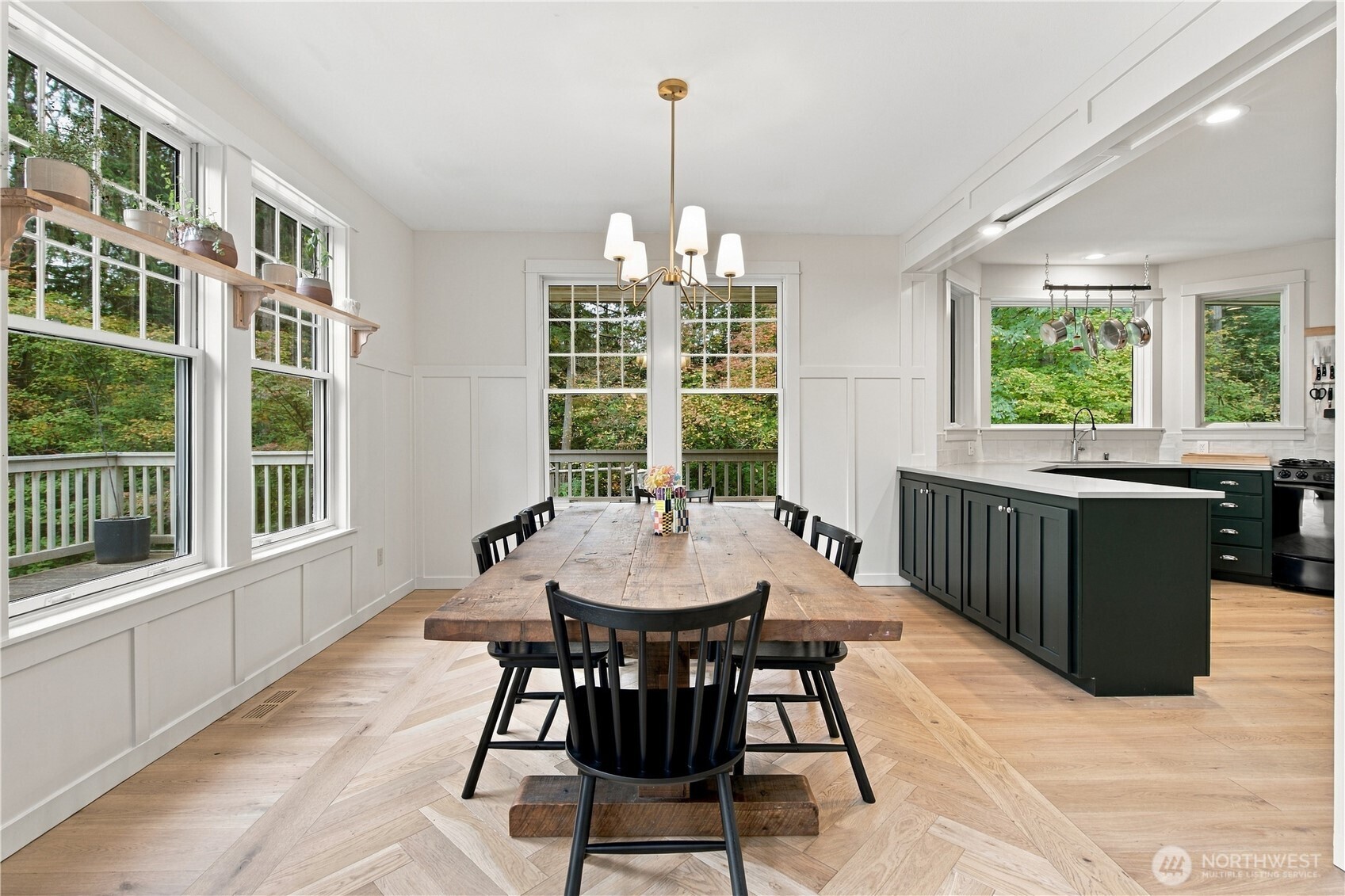 Dining area with a long rustic table beside large windows, open to a kitchen with dark green cabinets.