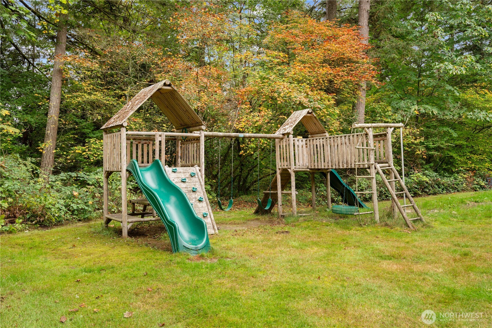 Wooden backyard playground with green slides and swings beneath autumn trees.