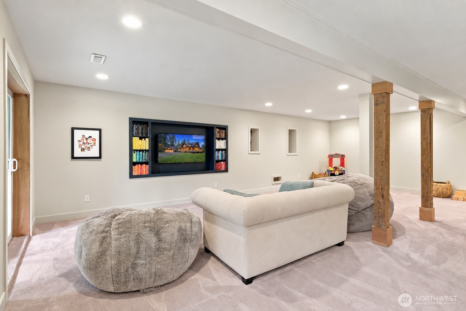 Lower-level family room with a built-in media wall, curved sofa, oversized ottomans, and exposed wooden posts.