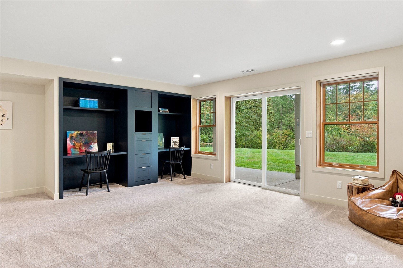 Home office with dark built-in desks and shelving, opening through a sliding glass door to a wooded lawn.
