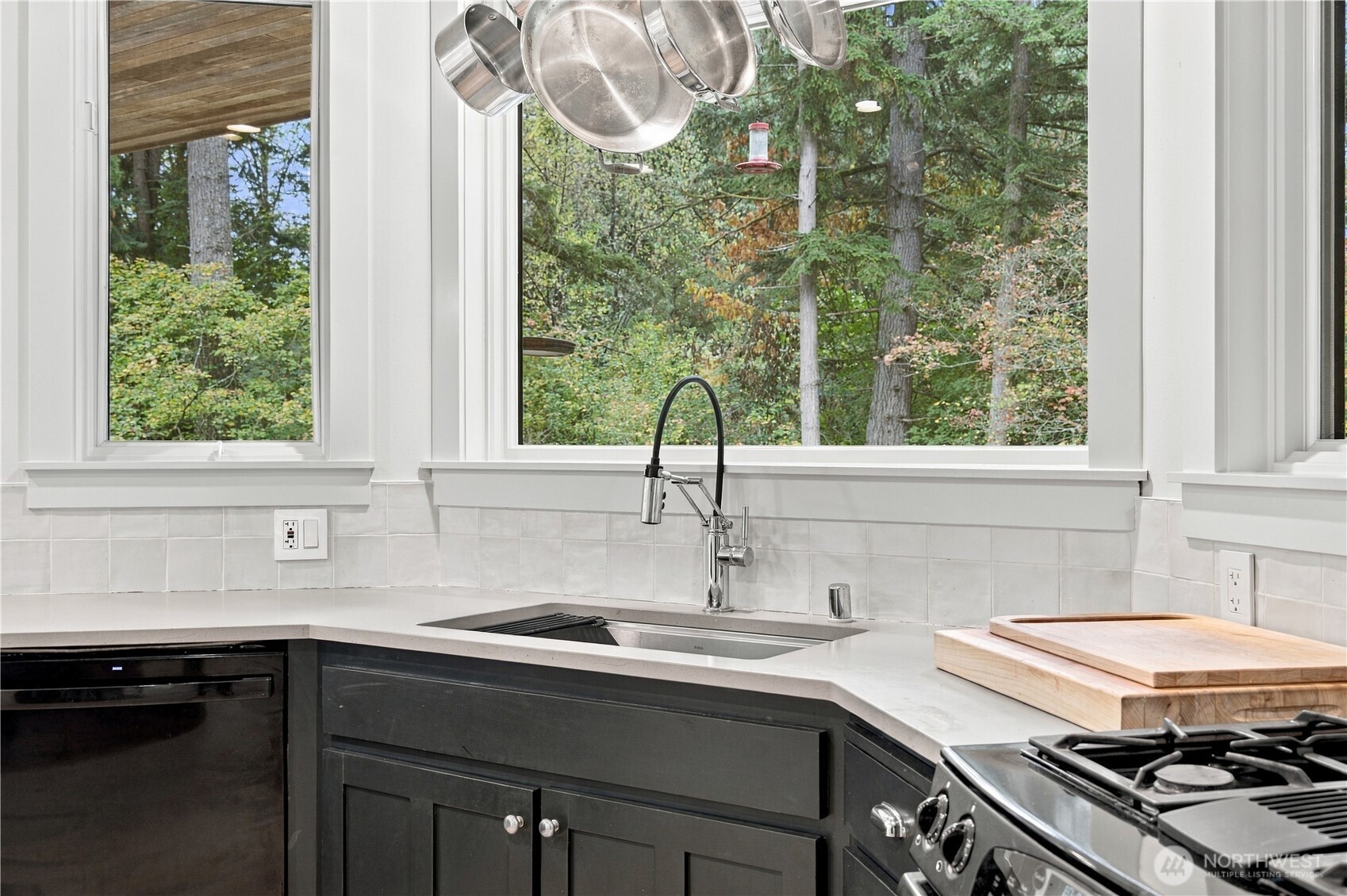 Kitchen sink beneath large windows overlooking trees, with dark cabinets, hanging pots, and a gas range.