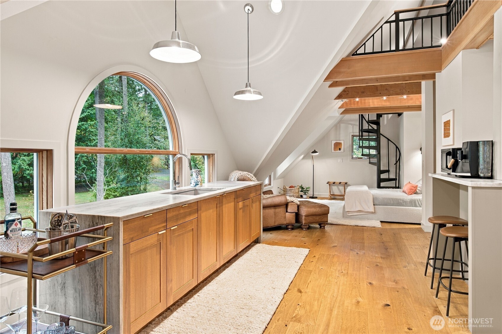 Open kitchen and living area with a marble island, arched woodland-view window, exposed beams, and spiral staircase.