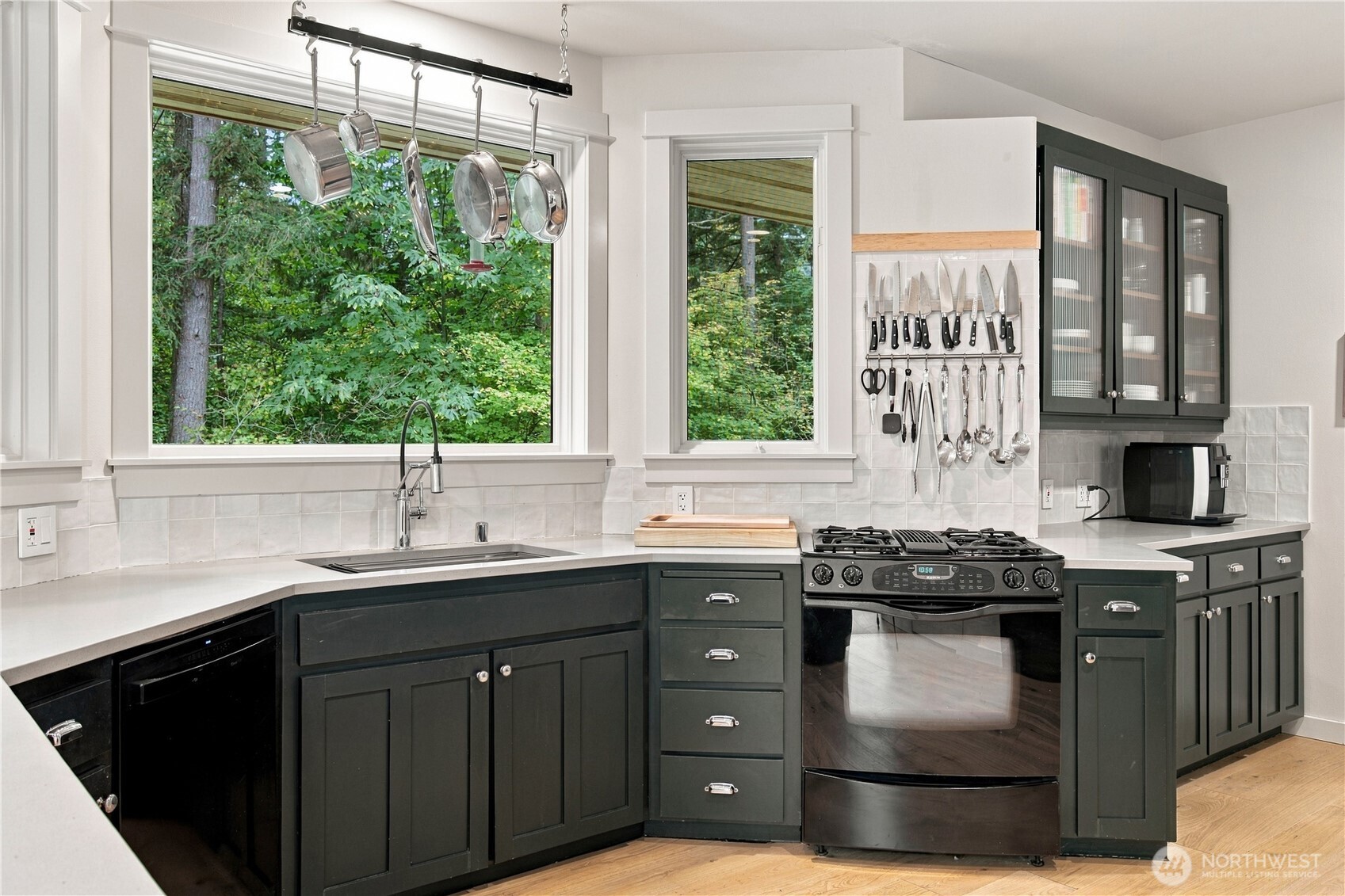 Kitchen with dark green cabinets, hanging cookware, and large windows overlooking trees.