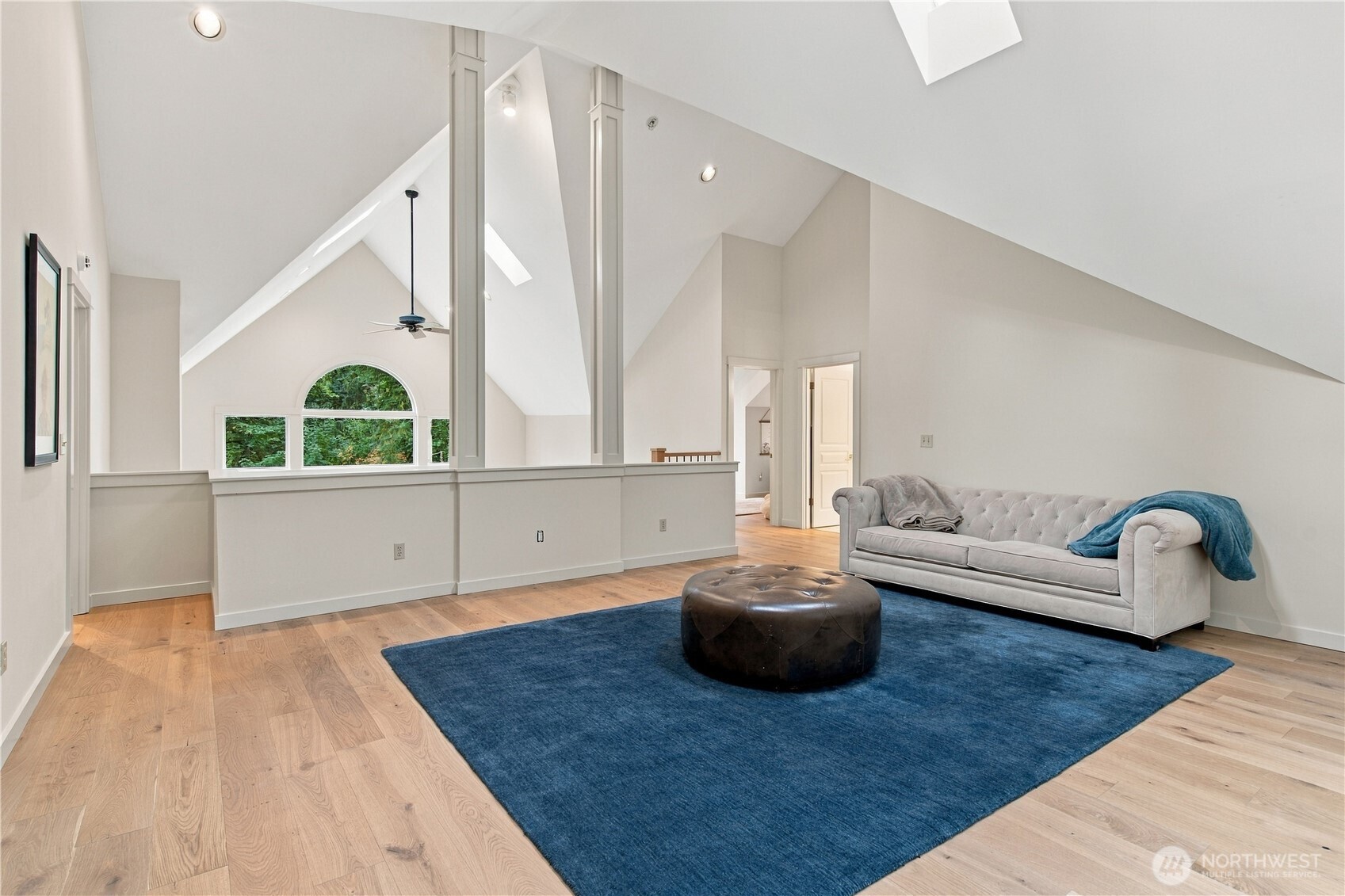 Vaulted living room with a gray sofa, blue rug, skylights, and arched window overlooking trees.