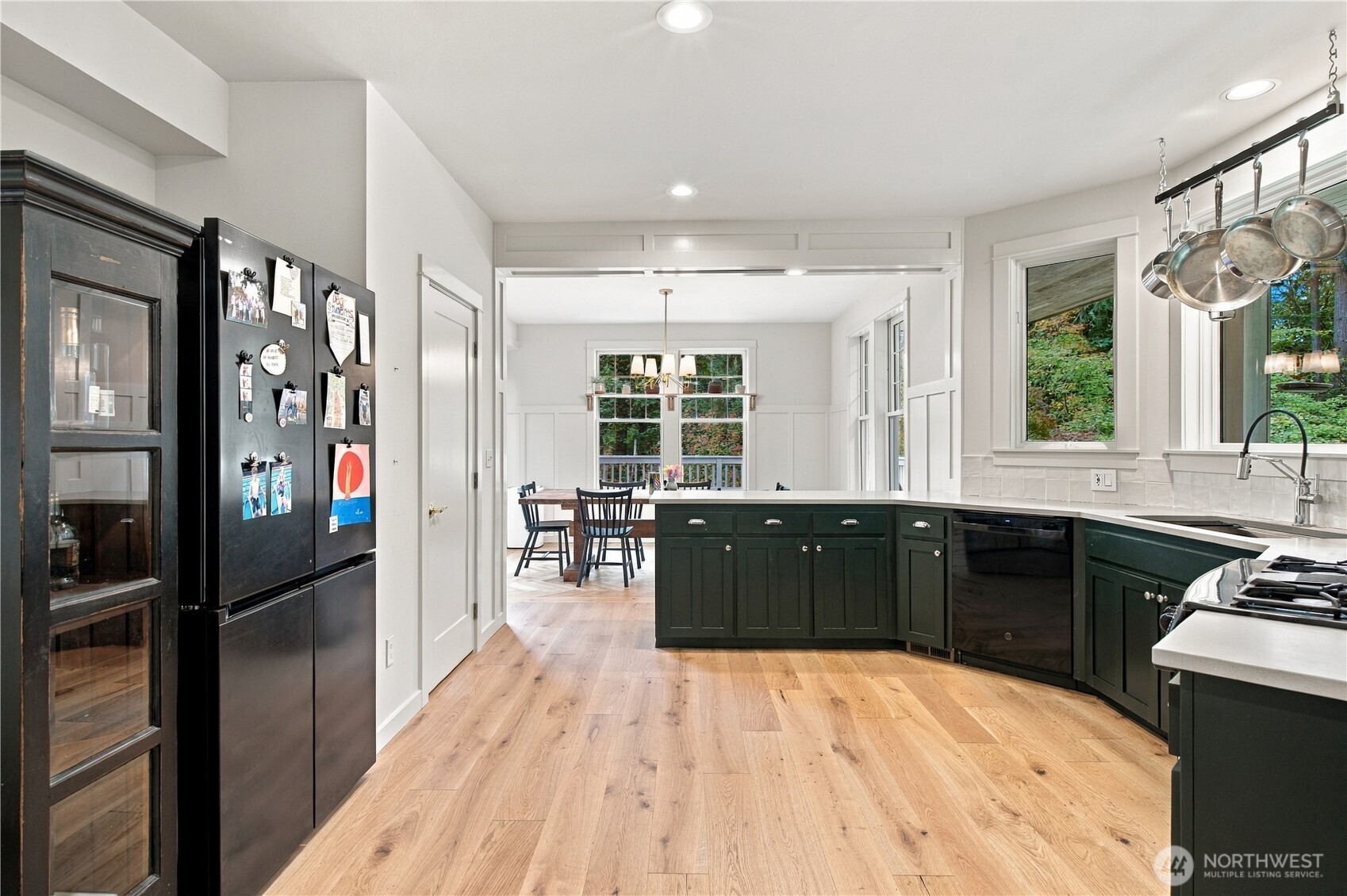 Kitchen with dark green cabinets, wide plank wood floors, and a dining area by large windows.